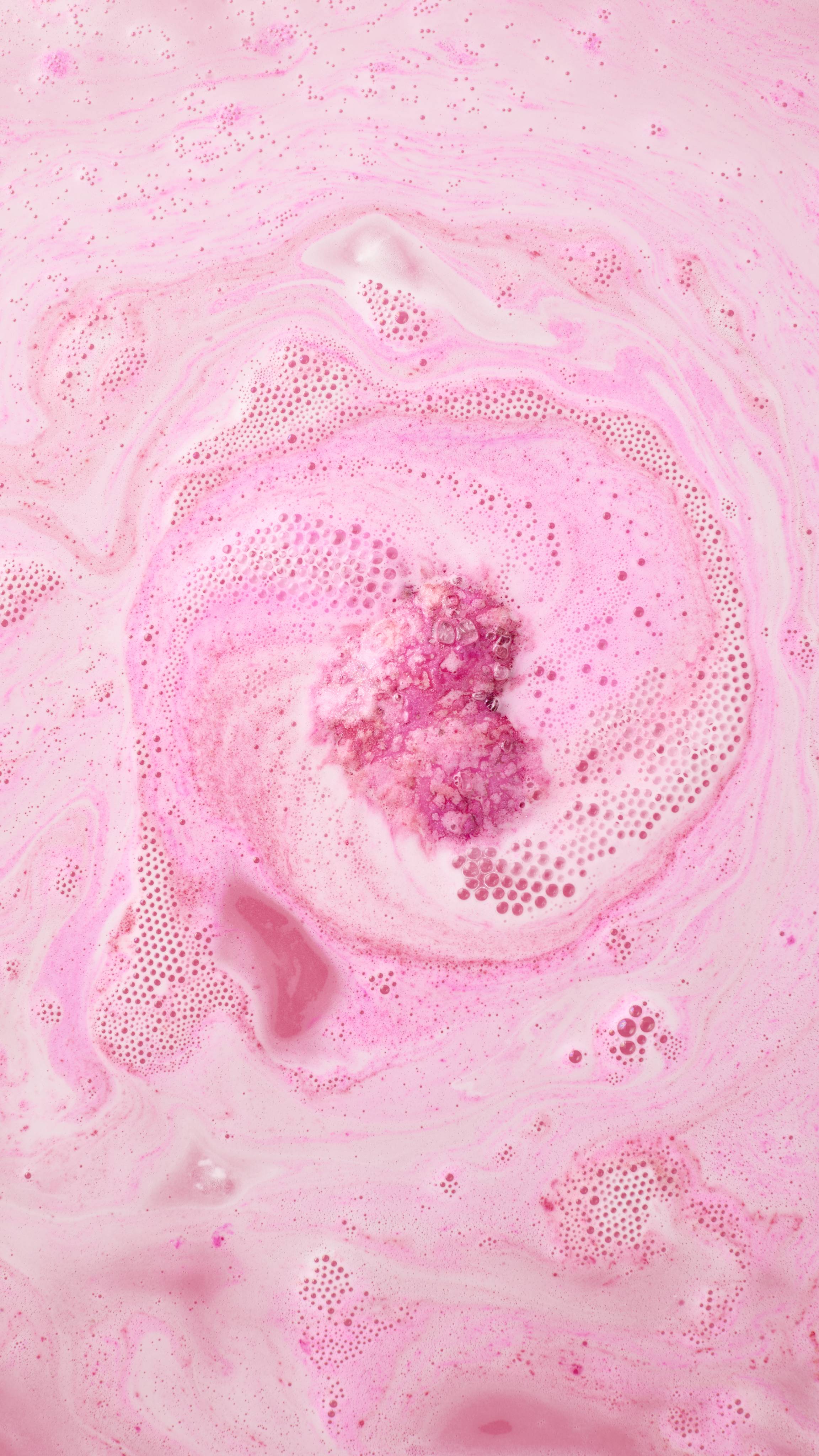 An aerial view of vibrant pink bathwater featuring thick, creamy white and dark pink swirls. Small clusters of bubbles and foam create a marbled pattern across the surface of the water.