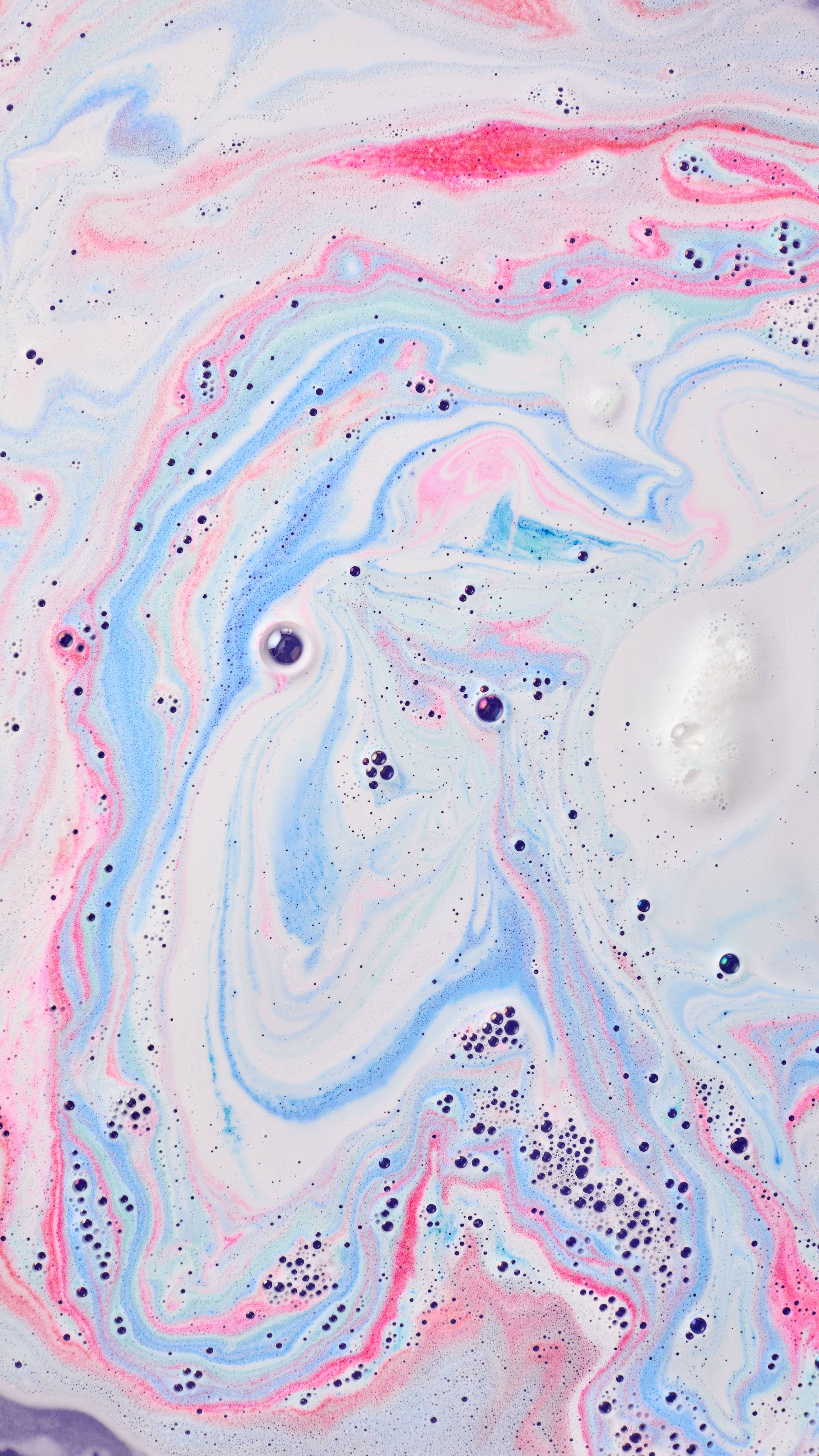 The image shows swirling bath art from the Mad Scientist Bath Bomb, a dreamy blend of pastel pink, light blue, and white hues. Intricate marbled patterns mix with scattered dark bubbles across the water surface.