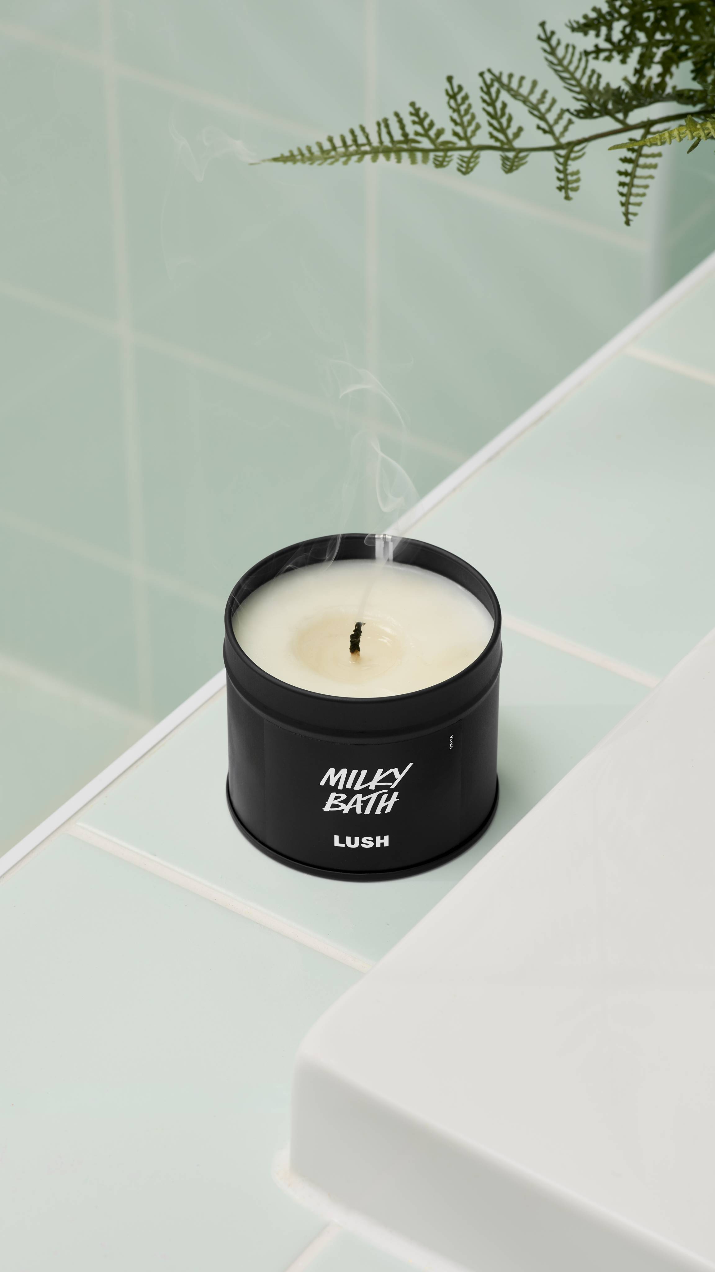 A close-up, high-angle shot of an extinguished Milky Bath candle by Lush in a matte black tin. Thin wisps of smoke rise from the blackened wick. It sits on a white ledge against mint green wall tiles with a fern leaf visible at the top.