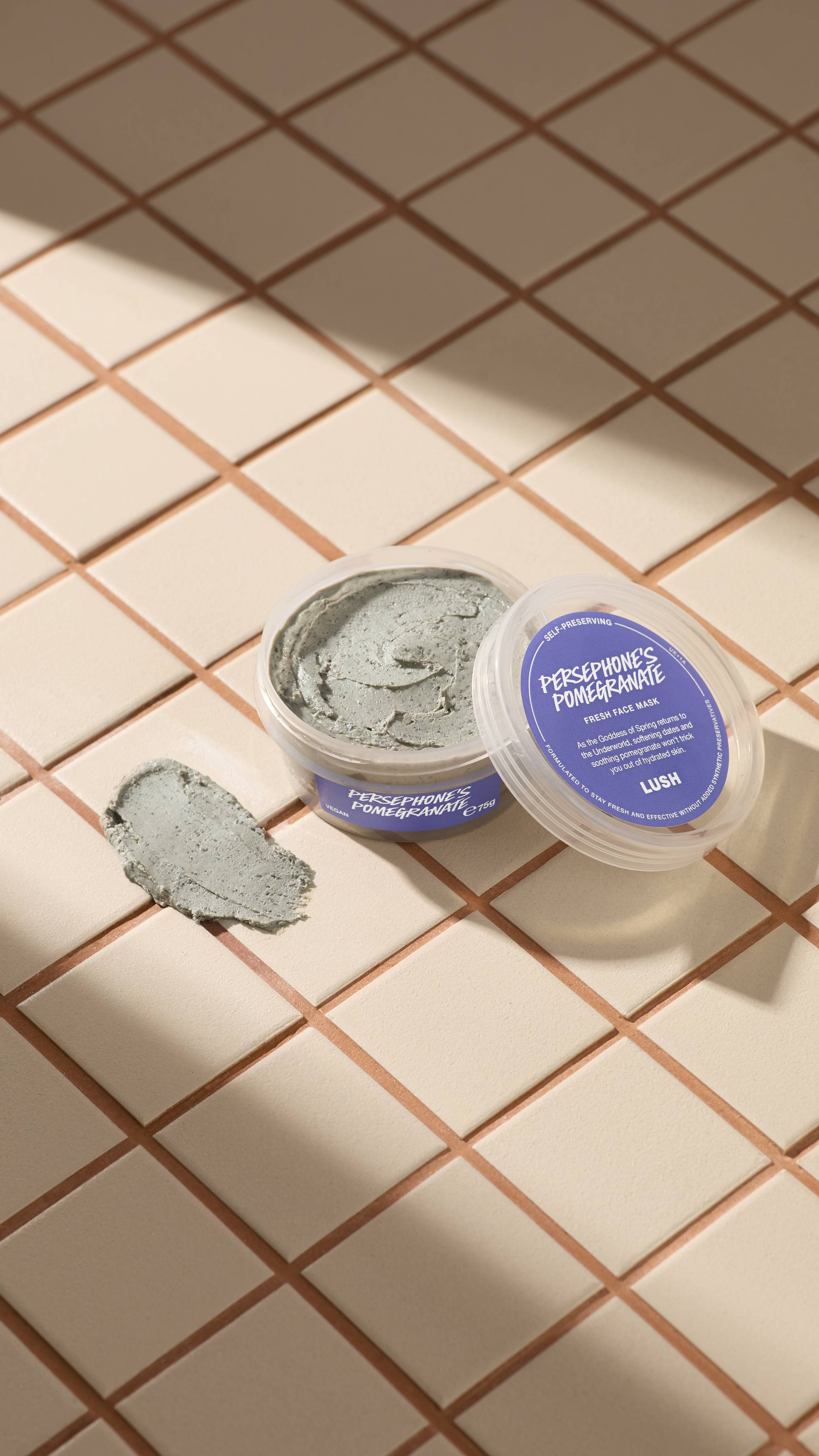 An open clear pot of Lush Persephone's Pomegranate fresh face mask sits on beige tiles next to a textured smear of the product. The clear lid with a purple label rests against the pot. Sunny shadows cross the tiled surface.