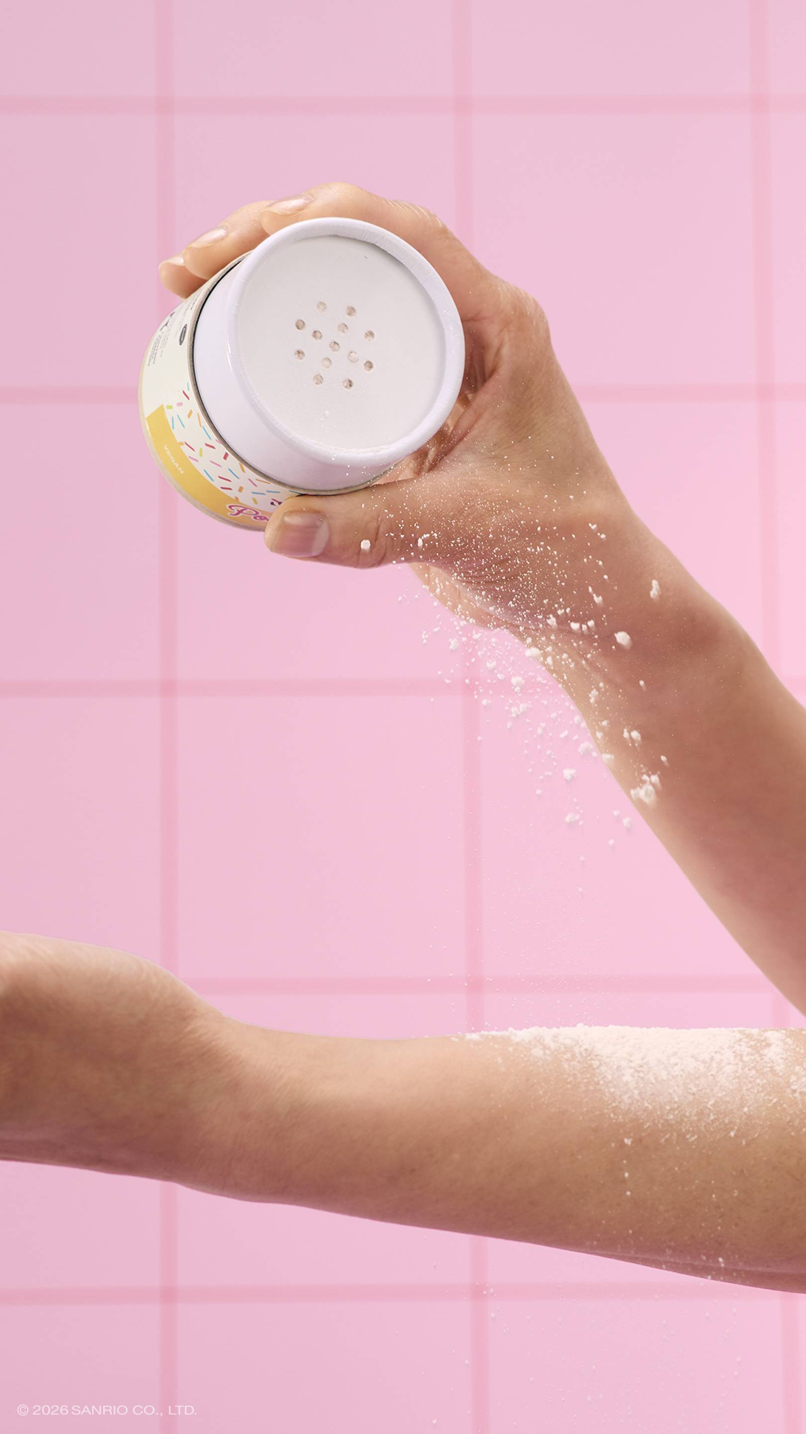 A close-up of a model's hand shaking white dusting powder from a Lush x Pochacco tube onto their other arm. The scene is set in a pink tiled bathroom.