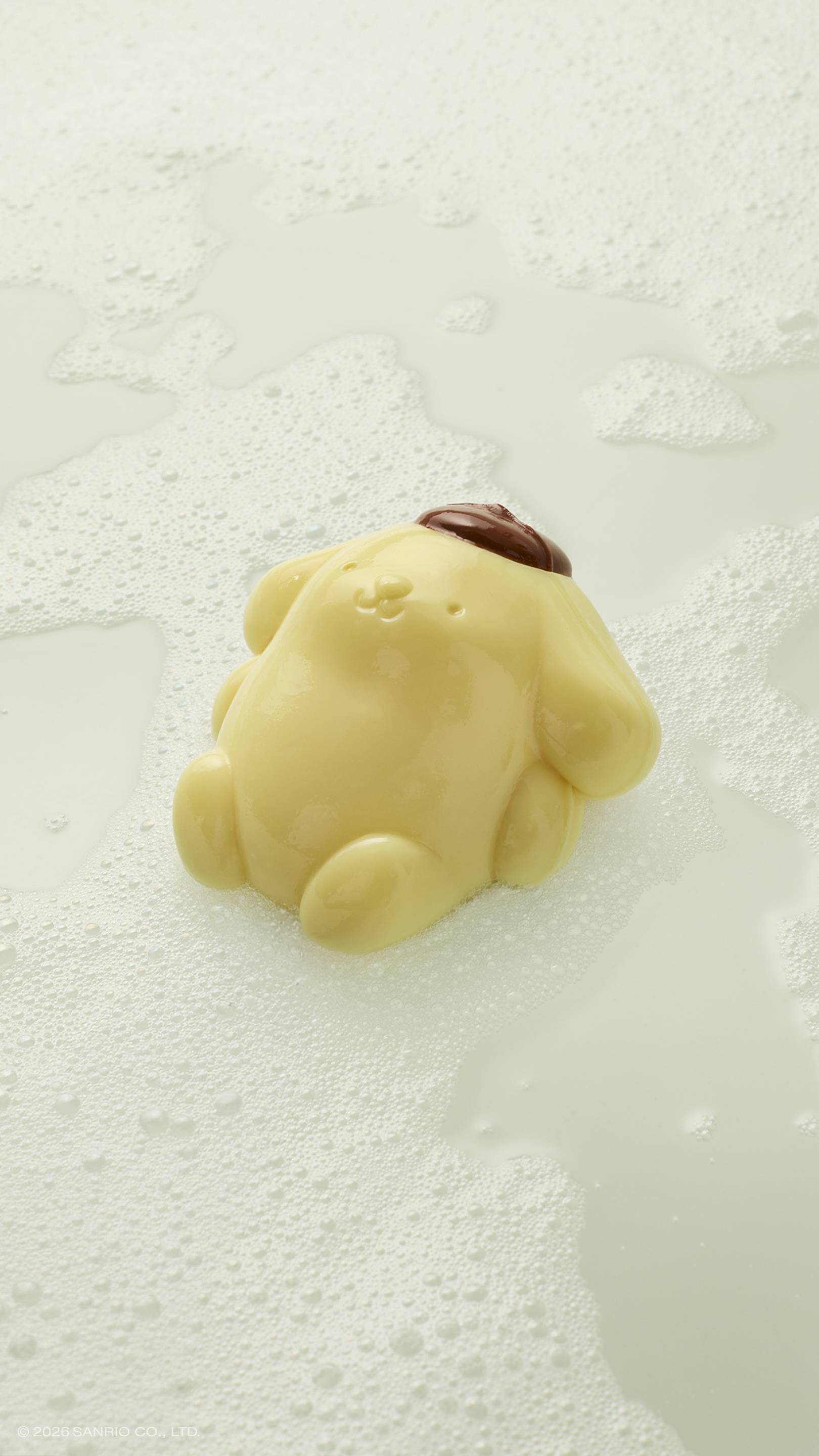 A glossy, pale yellow Pompompurin shower jelly rests in a pool of white soapy suds and bubbles. The character, wearing its signature brown beret, is shown from a high-angle view on a light, neutral surface.