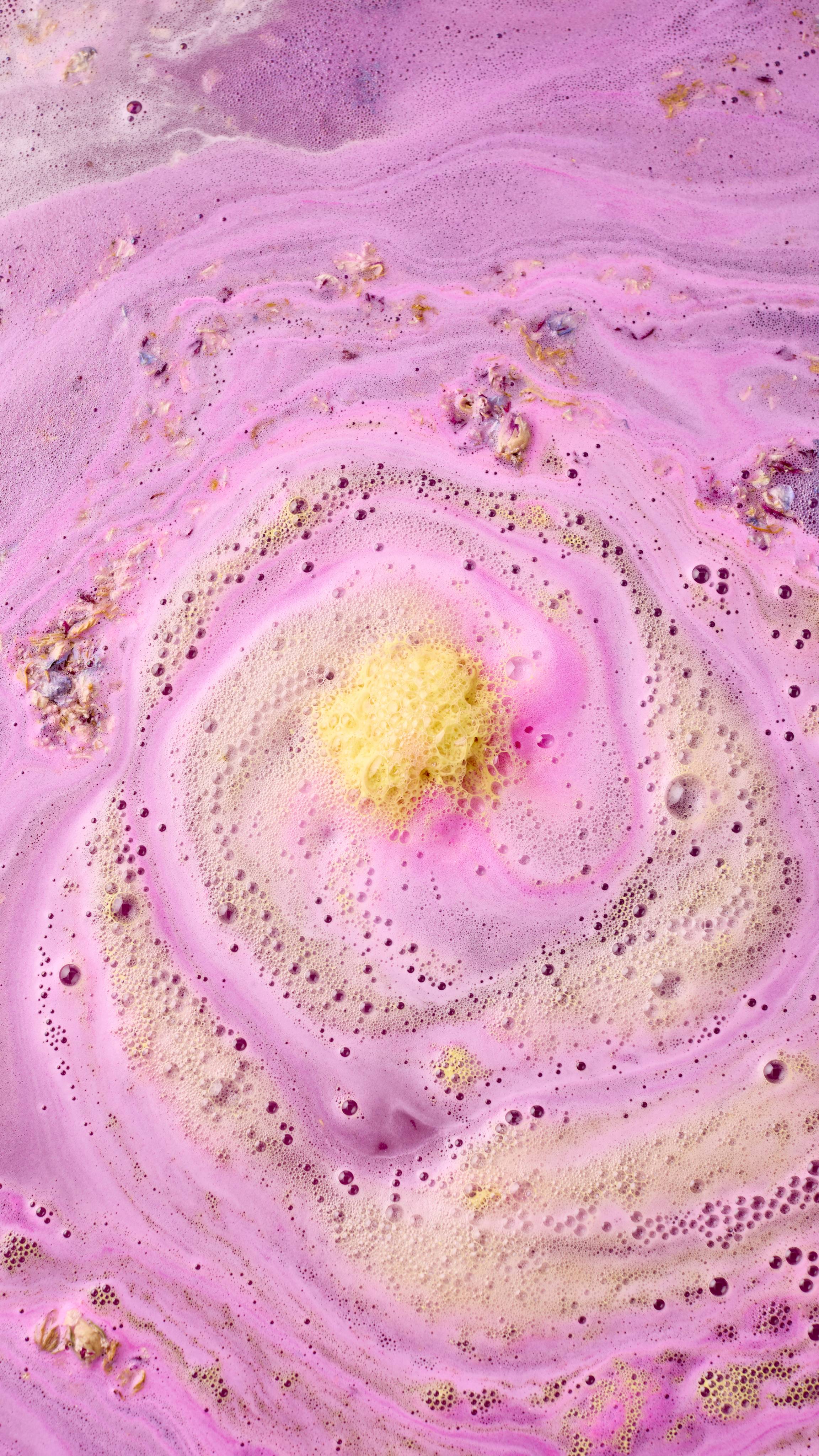 The bathwater swirls with vibrant pink and lilac hues, centred around a patch of frothy yellow foam. Dried flower petals float across the bubbly surface, creating a textured, floral bath art display.