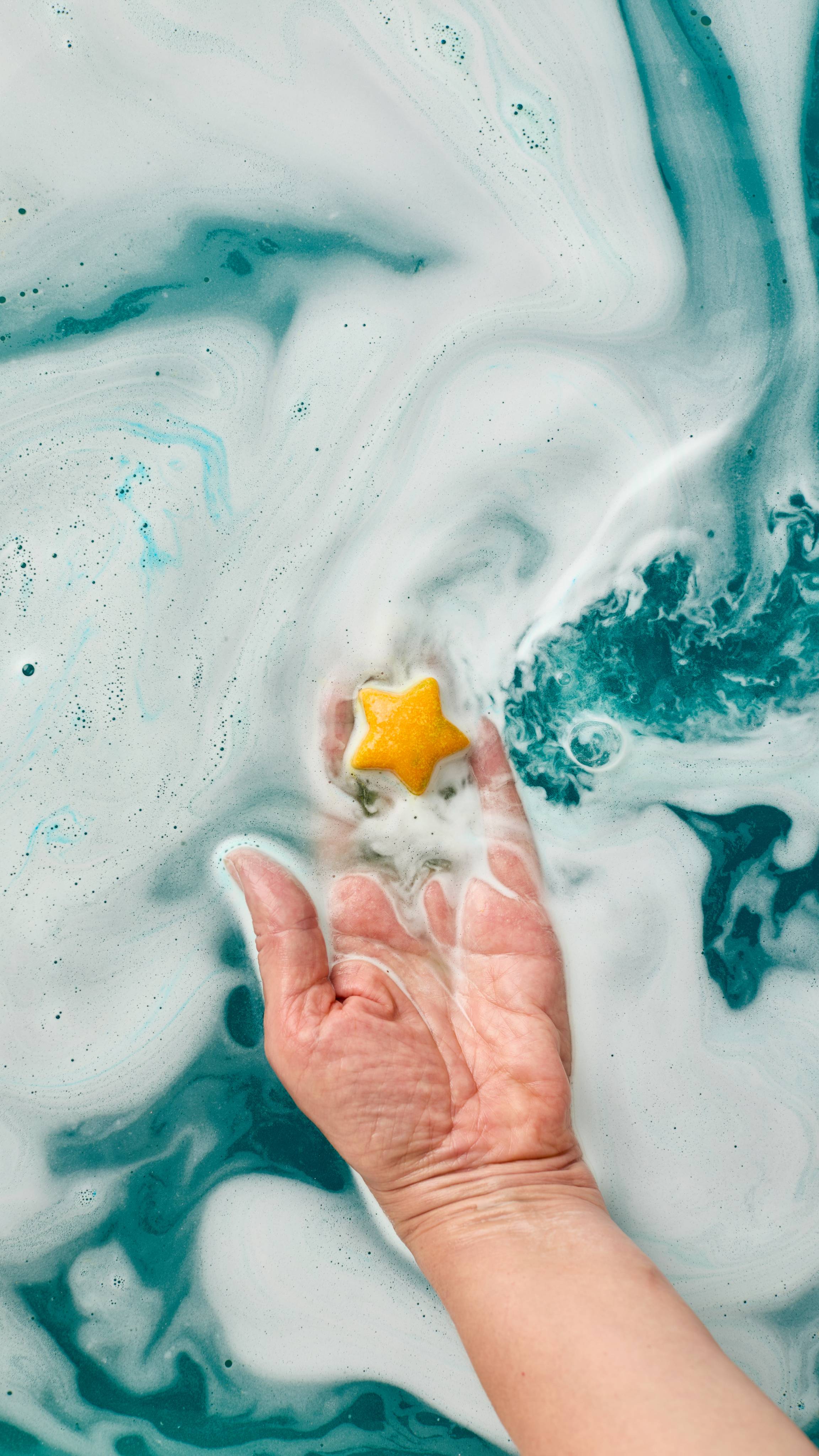 The image shows a hand cupping a golden, star-shaped bath melt from on top of the 'Shoot For The Stars' bath bomb after the rest of the product has dissolved. The product is held above swirling turquoise bath water with abundant white foam.