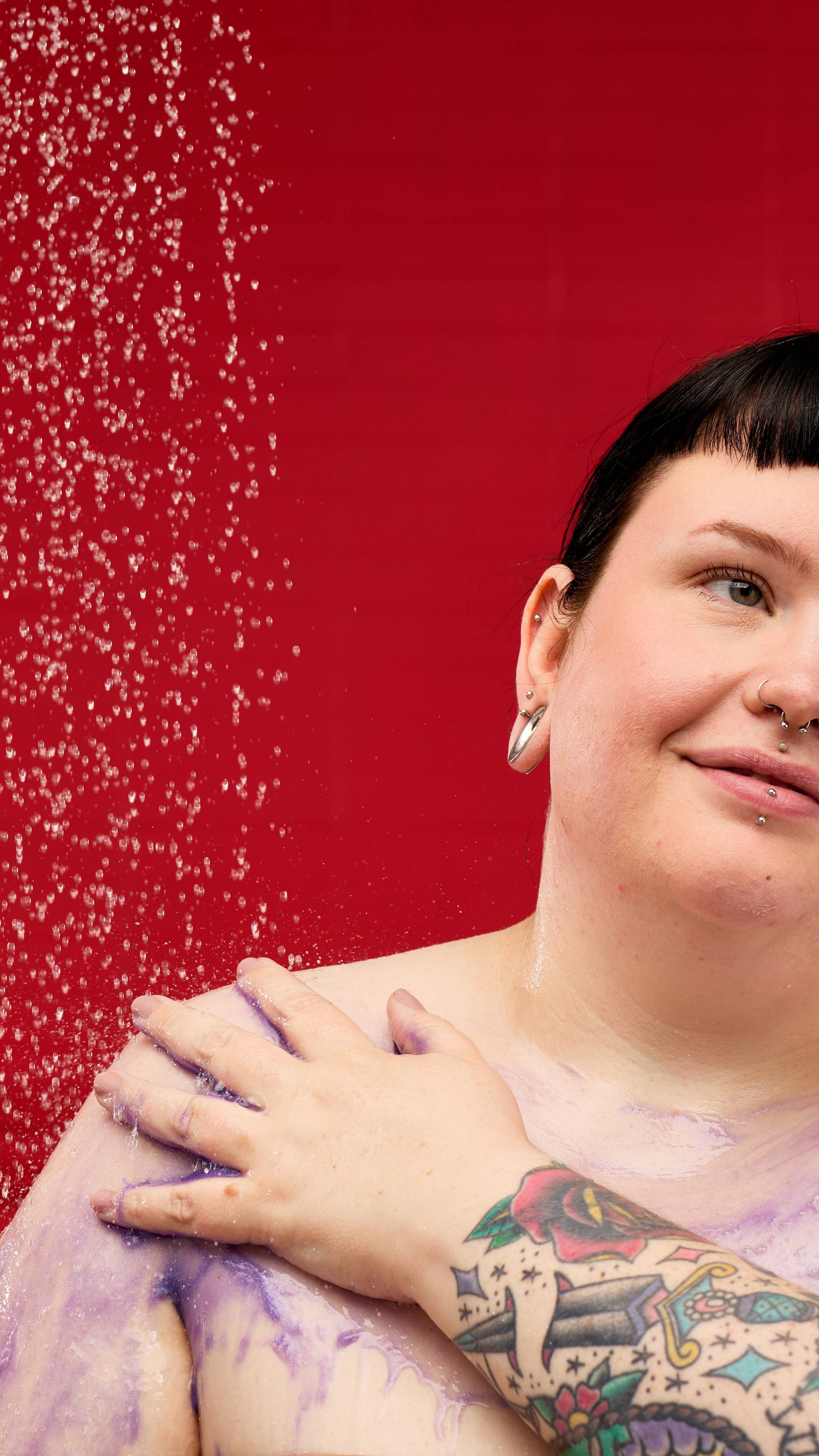 A person with a tattoo on their arm smiles while applying a purple, shimmering LUSH Sleepy Bear shower gel to their shoulder. Water droplets are visible in the air against a vibrant red background.