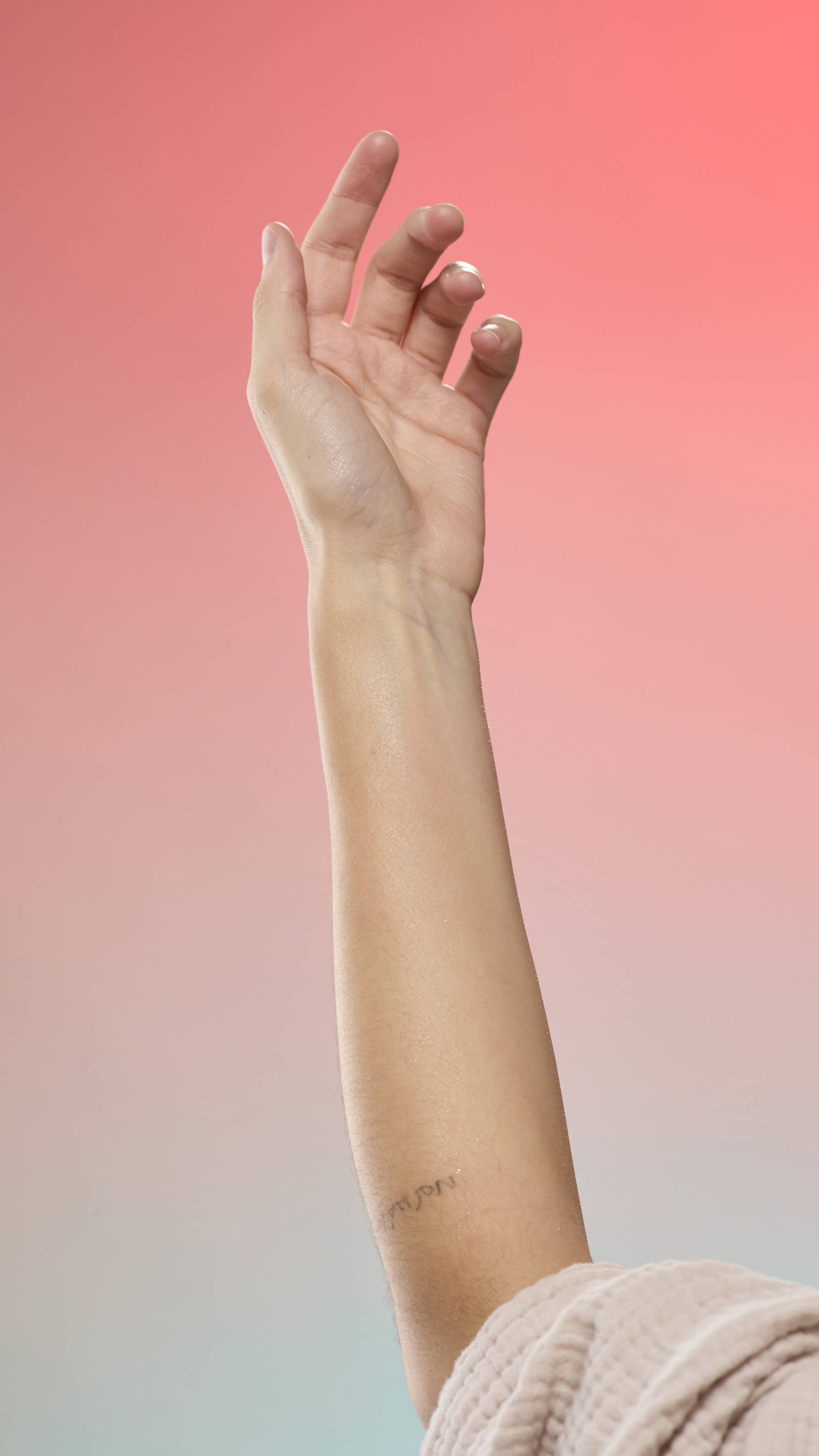 A tattooed arm shimmering with fine glitter against a peach-to-blue gradient background. The person is wearing a light, textured robe.
