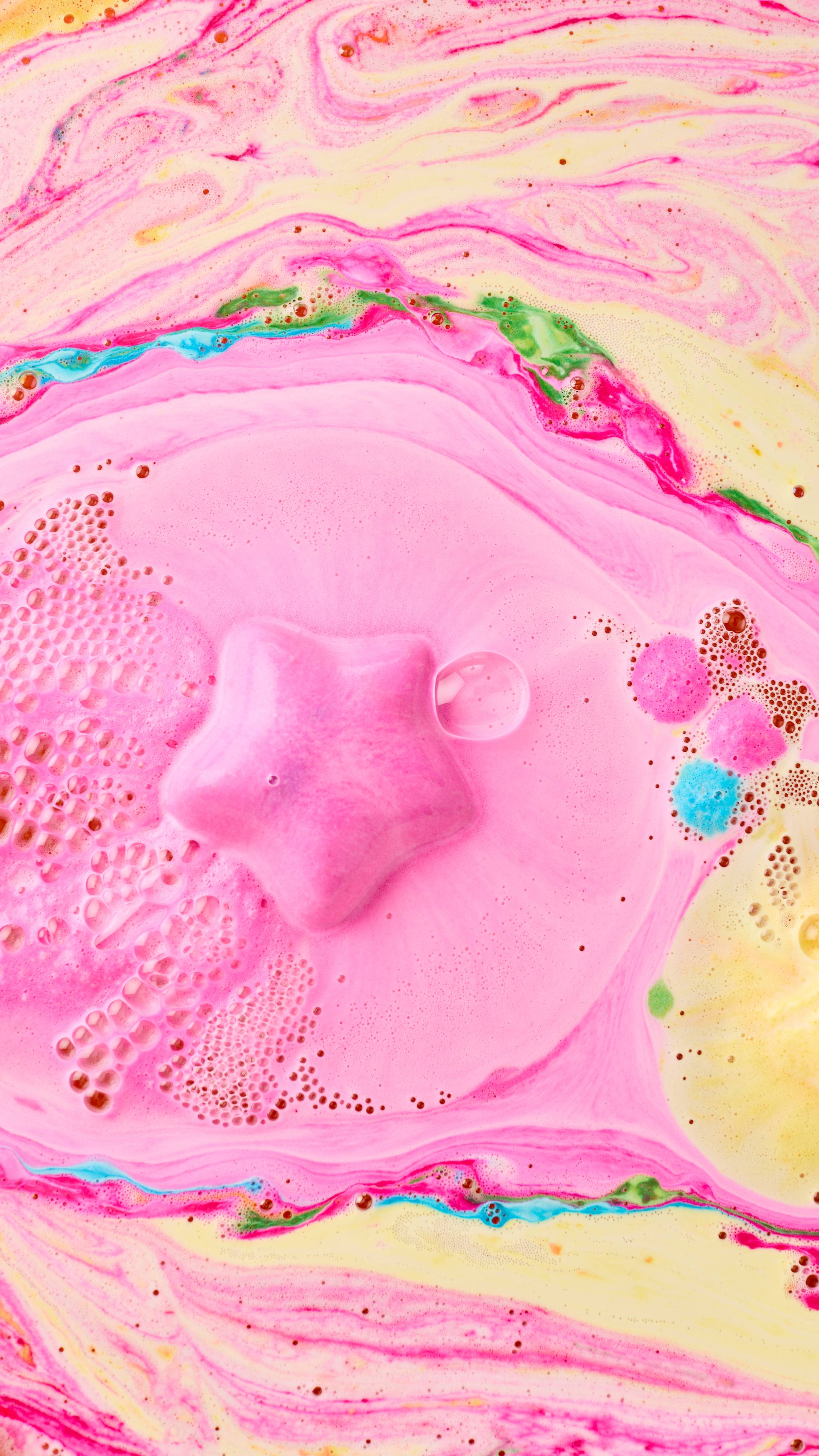 The image shows a vibrant pink Snow Fairy Lights bath bomb fizzing in swirling pink and yellow bath water. Pink, blue, and yellow hues blend with thick foam and bubbles.