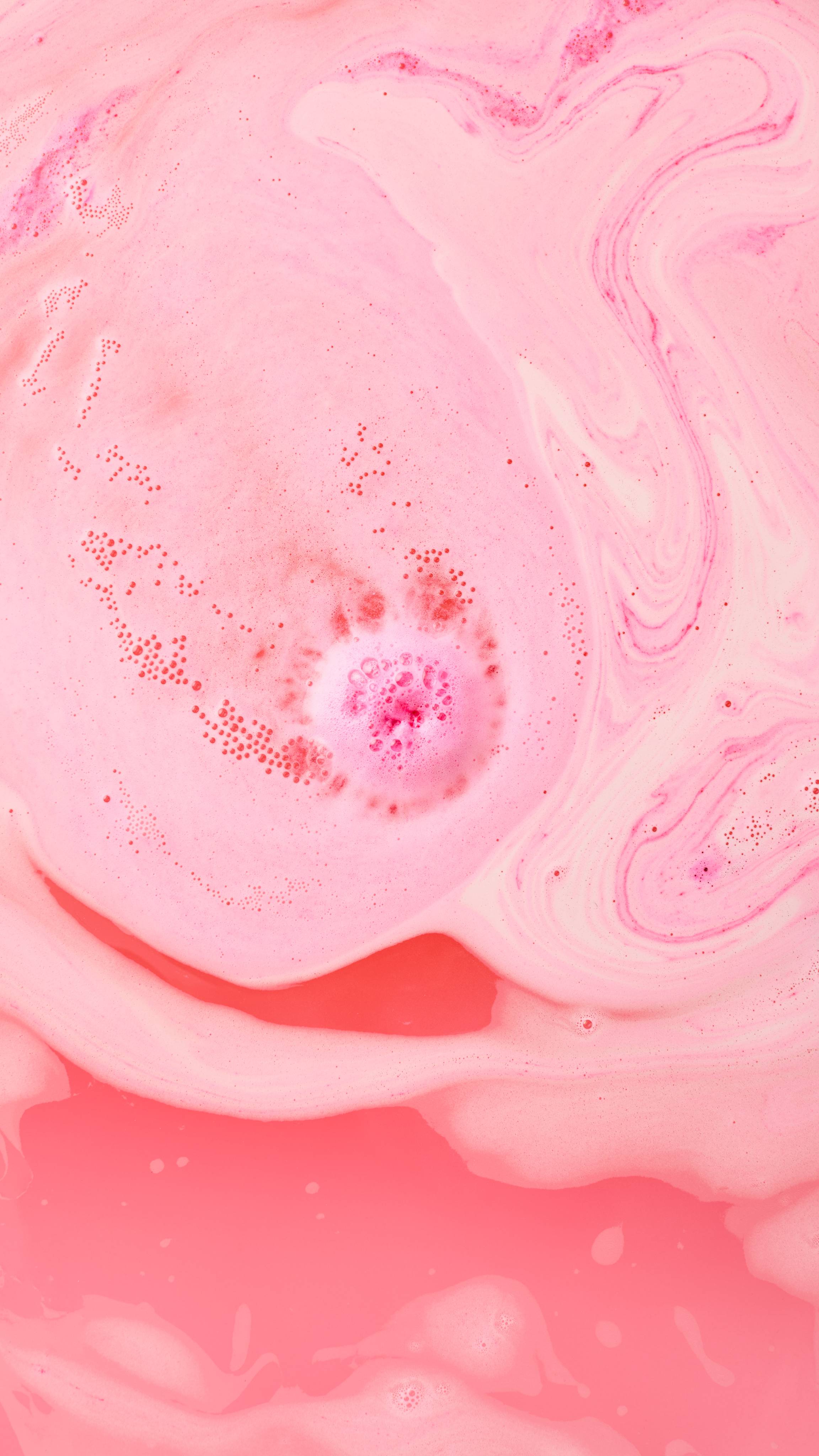 The image shows swirling pink bath water, with foam and light pink swirls. A concentrated core of red and dark pink bubbles is visible in the centre, where the Sweet Pudding bath bomb has recently dissolved.