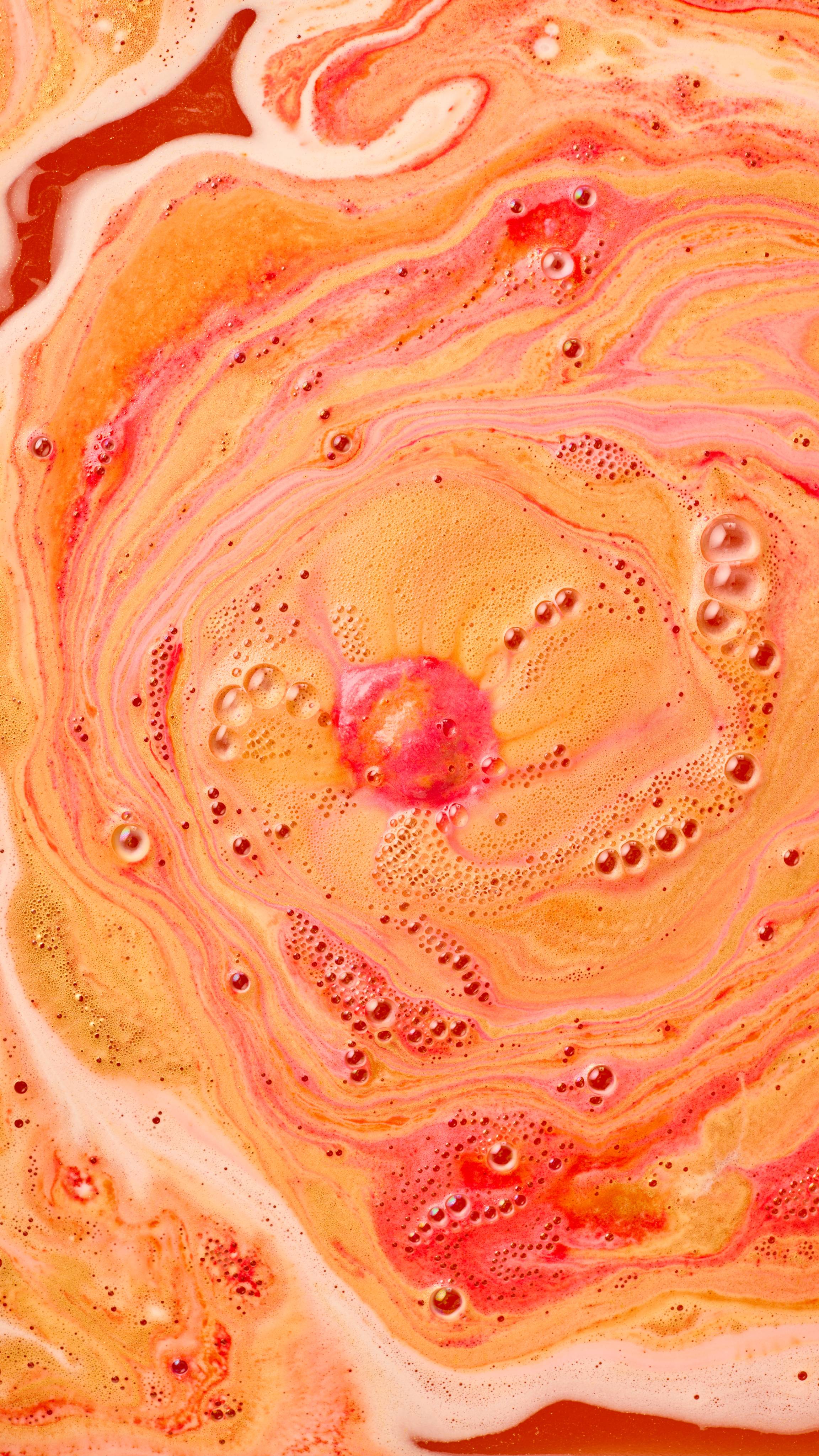 The image shows swirling bath art from 'The Alchemist' bath bomb, a fiery blend of vibrant orange, red-pink, and yellow. Intense, creamy swirls mix with prominent dark red bubbles and light foam across the water surface.