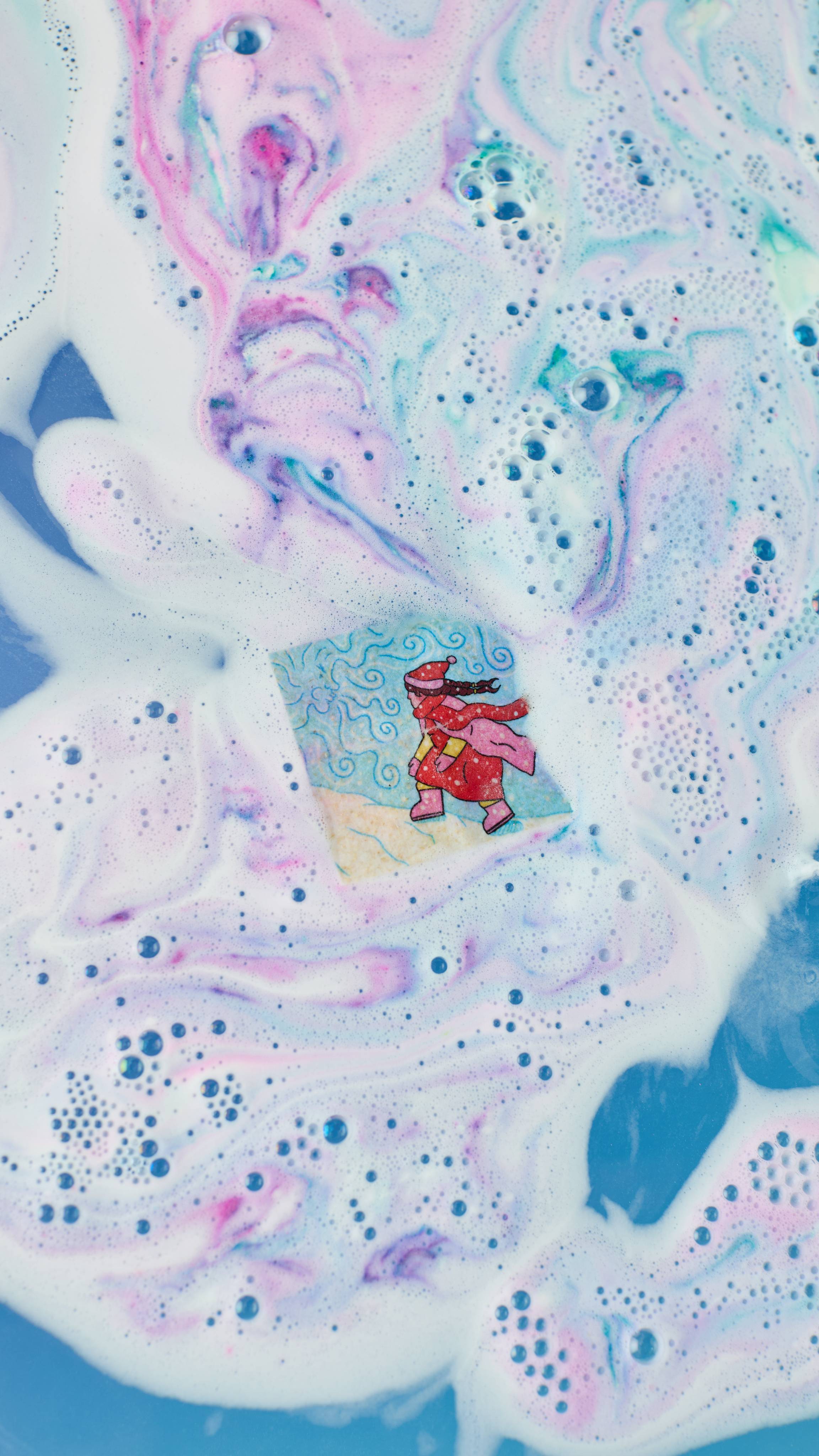 The image shows a small rice paper sheet with a cartoon of a person in a red coat, dissolving in swirling bath water. Pink, blue, and purple streaks mix with white foam and bubbles around the paper. 