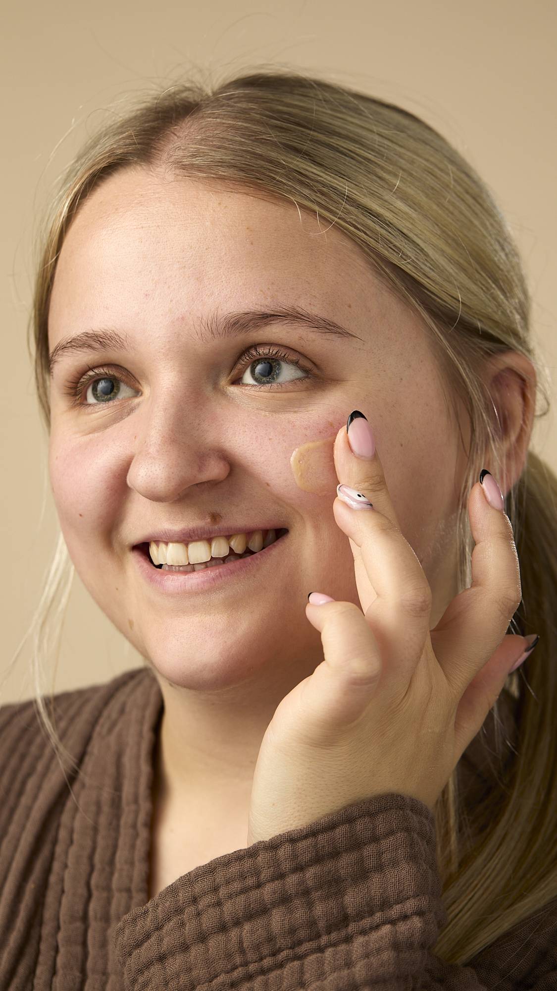 The image shows a light-skinned model with blonde hair pulled back, smiling while applying the Vision facial serum to the cheek under their right eye. They're wearing a brown, textured robe on a light beige background.