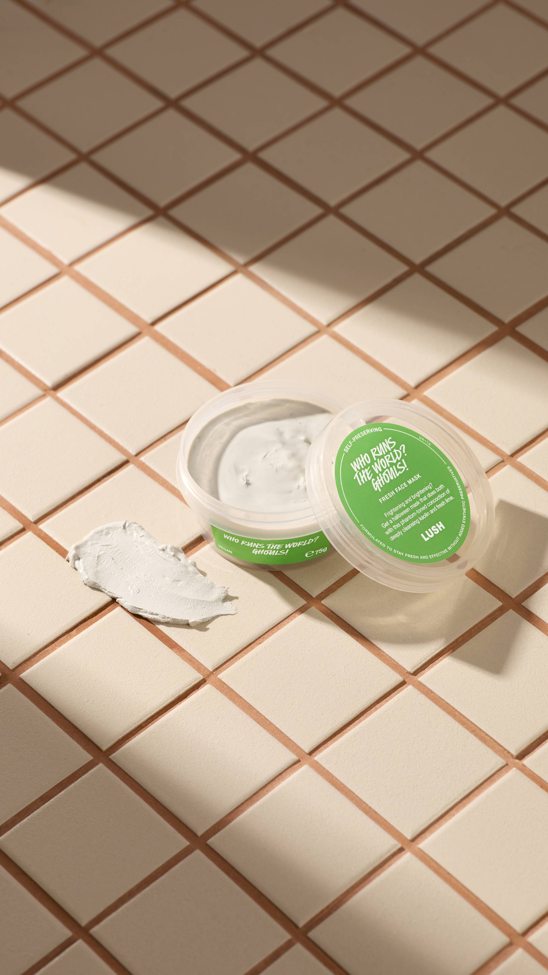 An open clear pot of Lush Who Runs The World? Ghouls! fresh face mask sits on beige tiles next to a textured smear of the product. The clear lid with a green label rests against the pot. Sunny shadows cross the tiled surface.