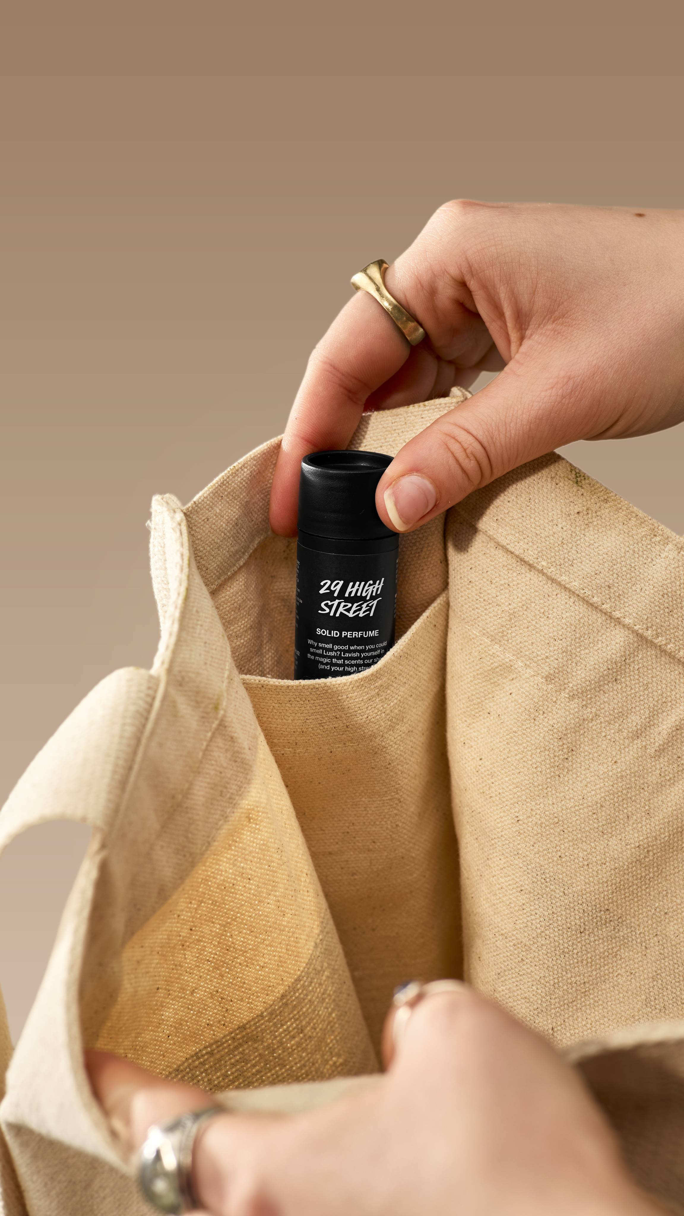 A close-up shot shows a hand placing a small, black Lush '29 High Street' solid perfume tube into the inner pocket of a cream canvas tote bag. The person is wearing several rings, set against a neutral beige background.