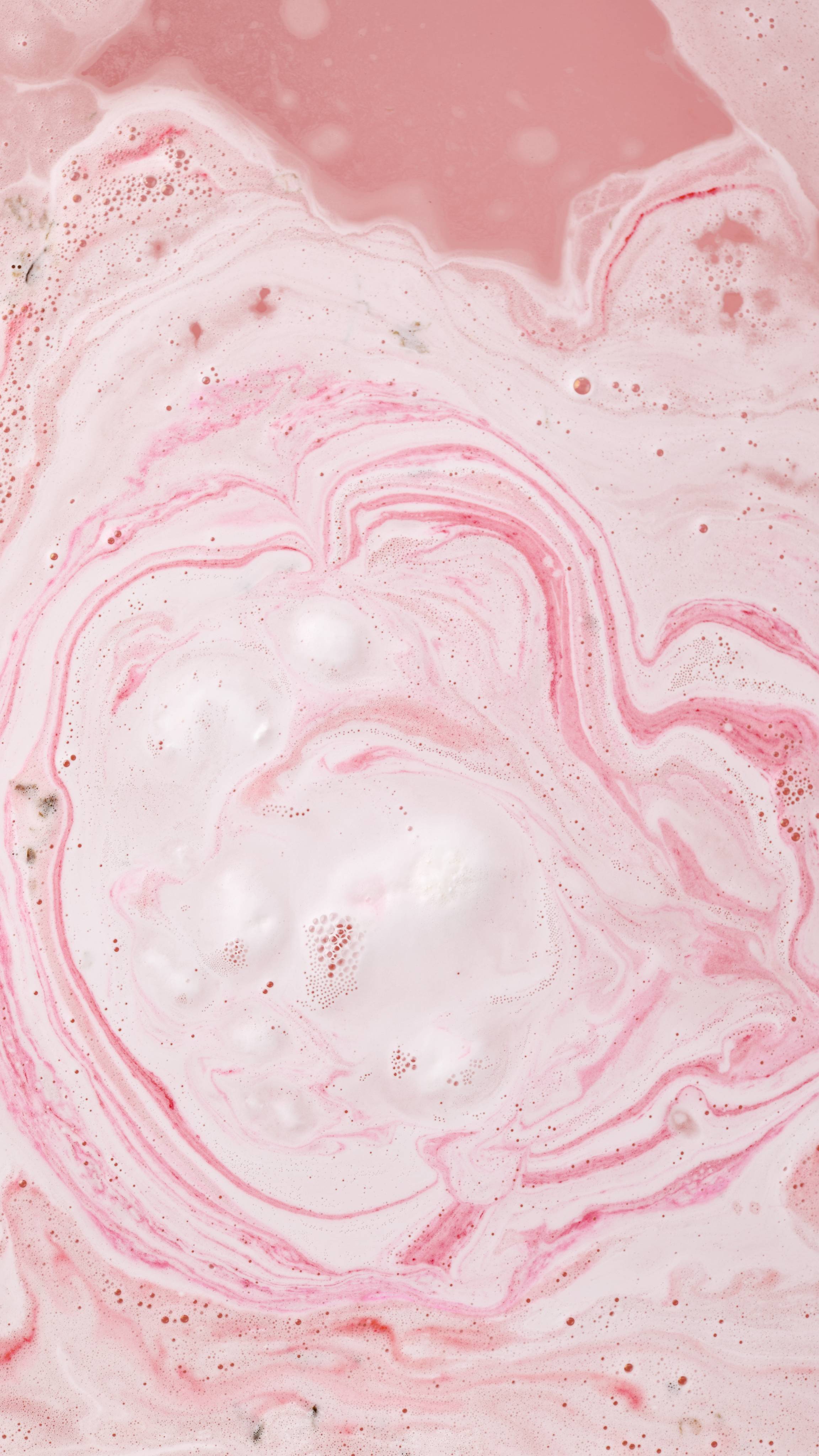 An aerial view of the bath after using the Blooming Cherry Blossoms bath bomb. There are swirling patterns of pale pink foam and vibrant magenta ripples and a marbled texture across the surface, with a dense white frothy area in the centre.