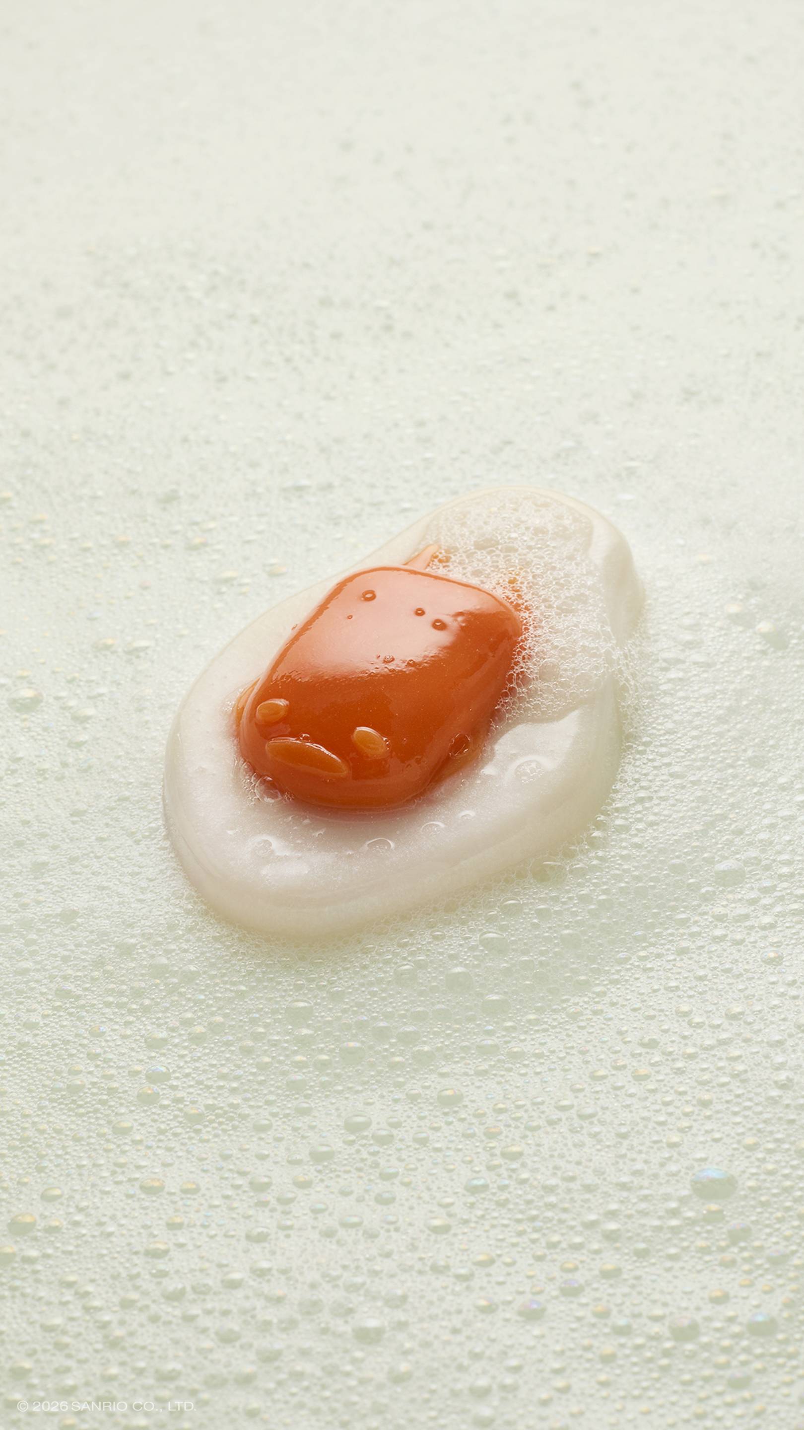 A glossy, fried-egg-shaped shower gummy rests in a pool of white soapy suds and bubbles. The orange "yolk" in the centre features the faint, sleepy face of the character Gudetama. 