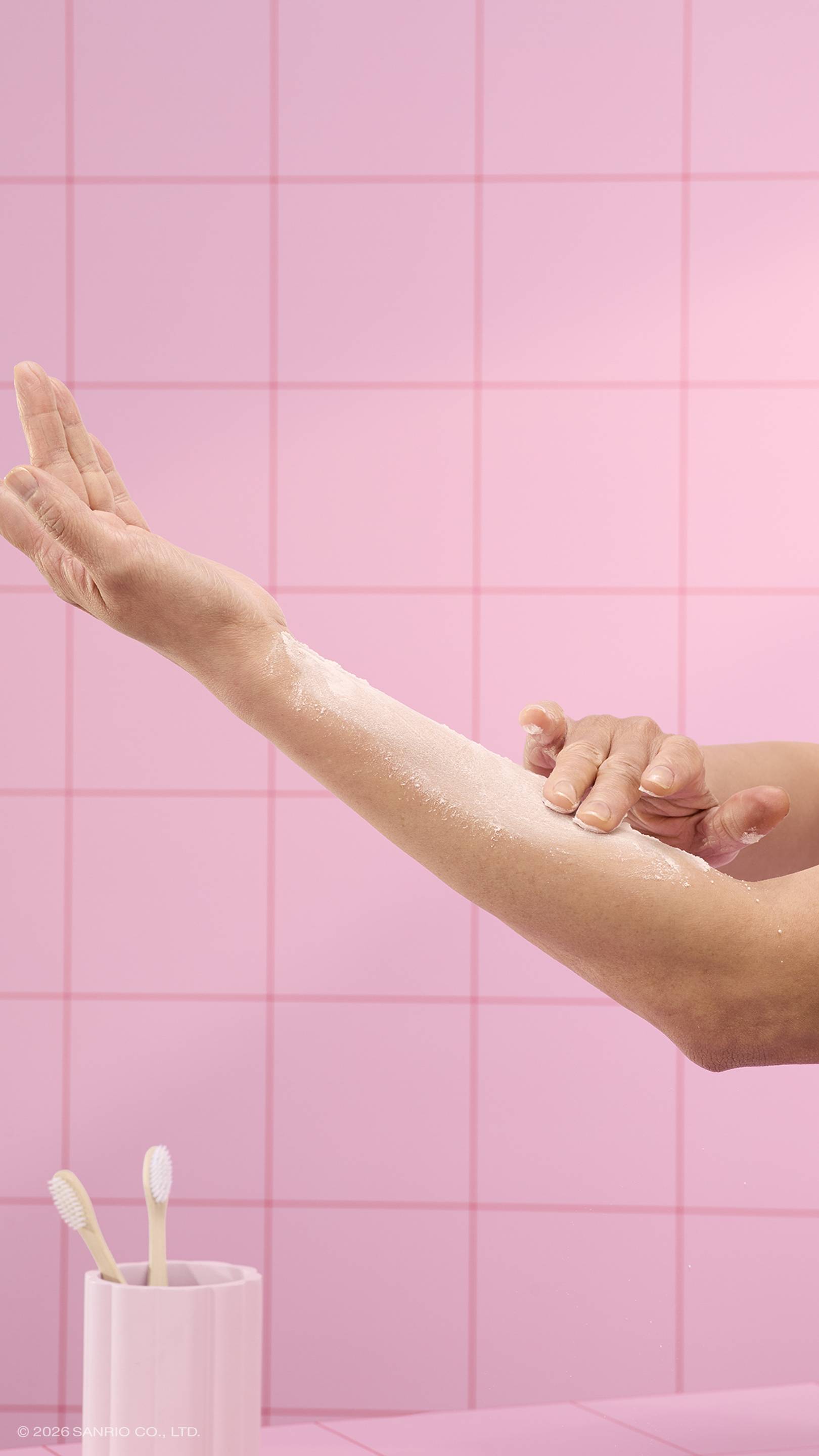 A close-up shows hands gently rubbing white dusting powder into an arm. The scene is set against a background of pink square tiles, with a pink toothbrush holder containing two toothbrushes in the lower-left corner. 