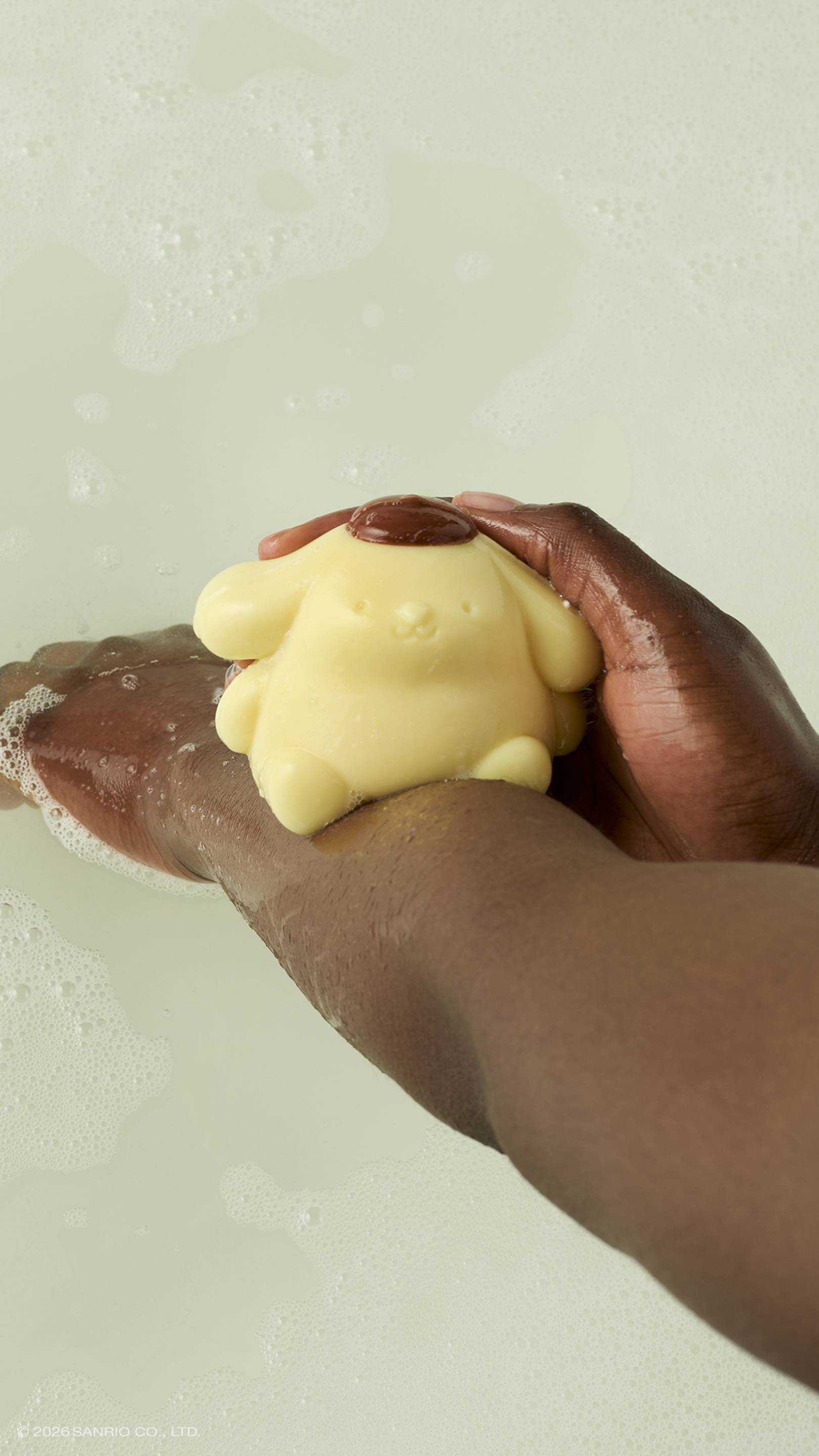 A model rubs a pale yellow Pompompurin shower jelly character against their arm. The glossy, dog-shaped character has a small brown beret and is held over a bath with soft bubbles on the surface.