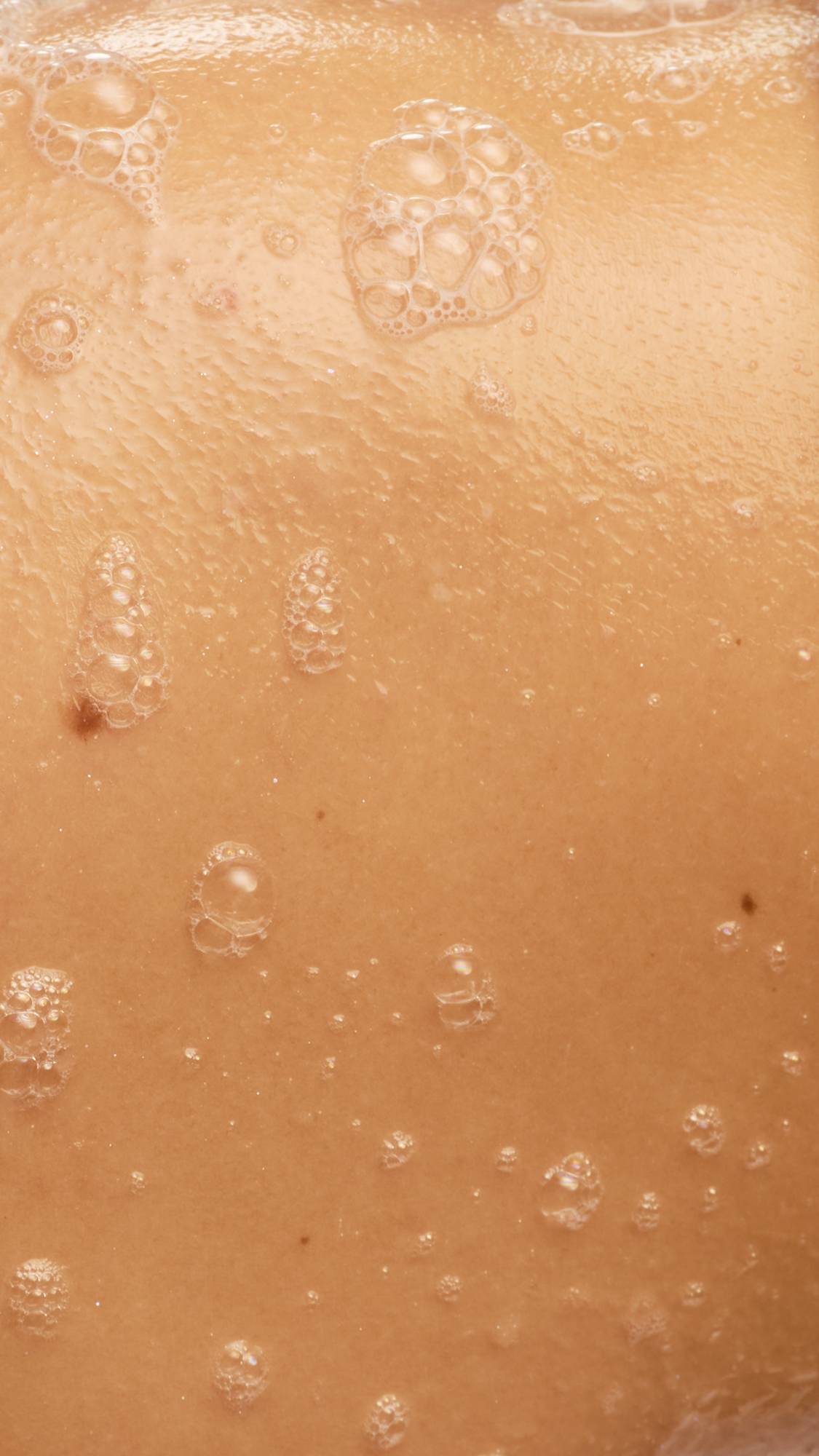 A close-up of wet skin covered in a light, bubbly lather after using Lush "Sex Bomb" shower gel. Small, translucent soap bubbles are scattered across the surface, reflecting light against the warm skin tone.