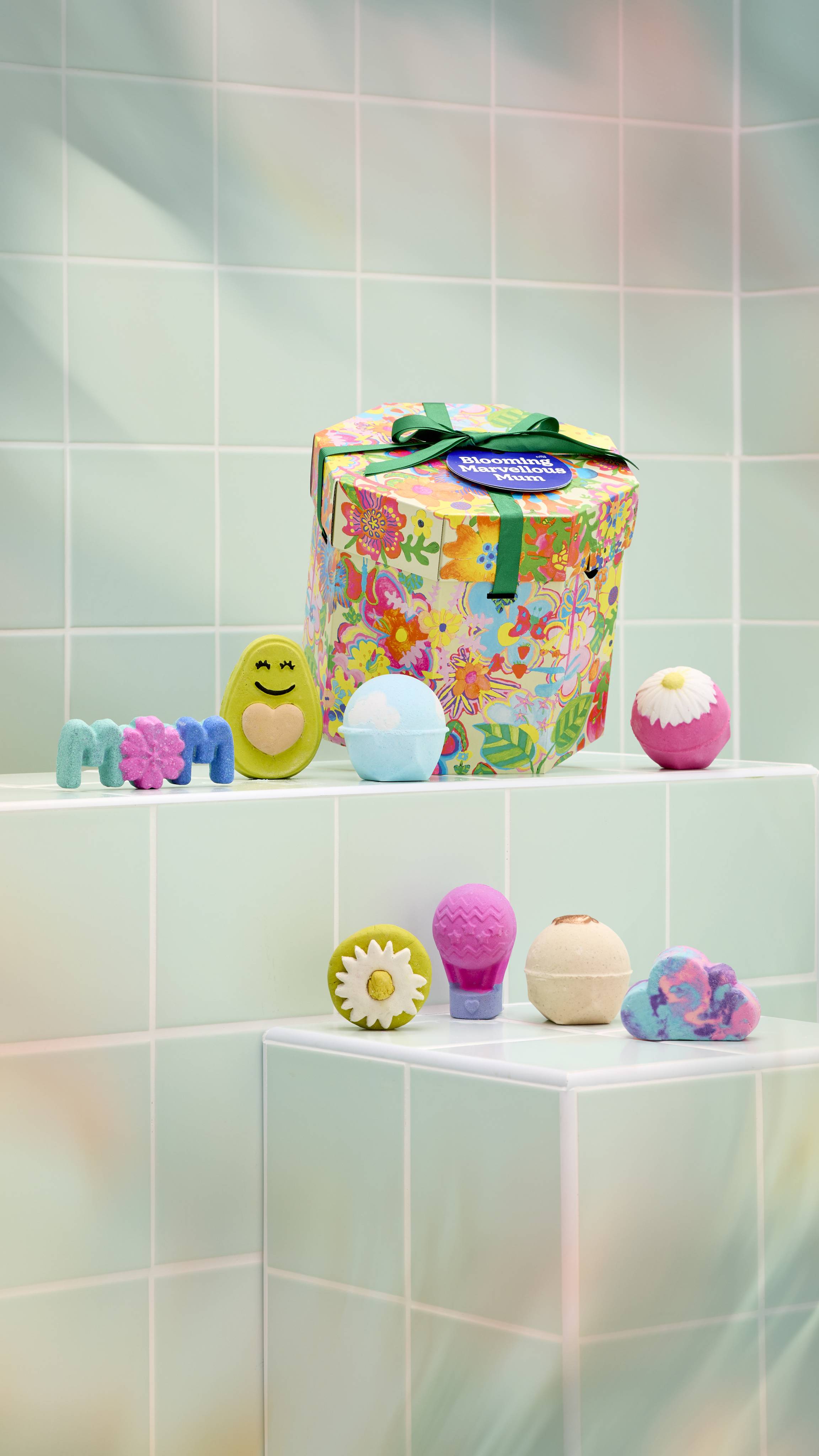 A vibrant, hexagonal "Blooming Marvellous Mum" gift box with a floral pattern sits on a tiled ledge. Arranged in front are various bath products, including five bath bombs and three bubble bars.