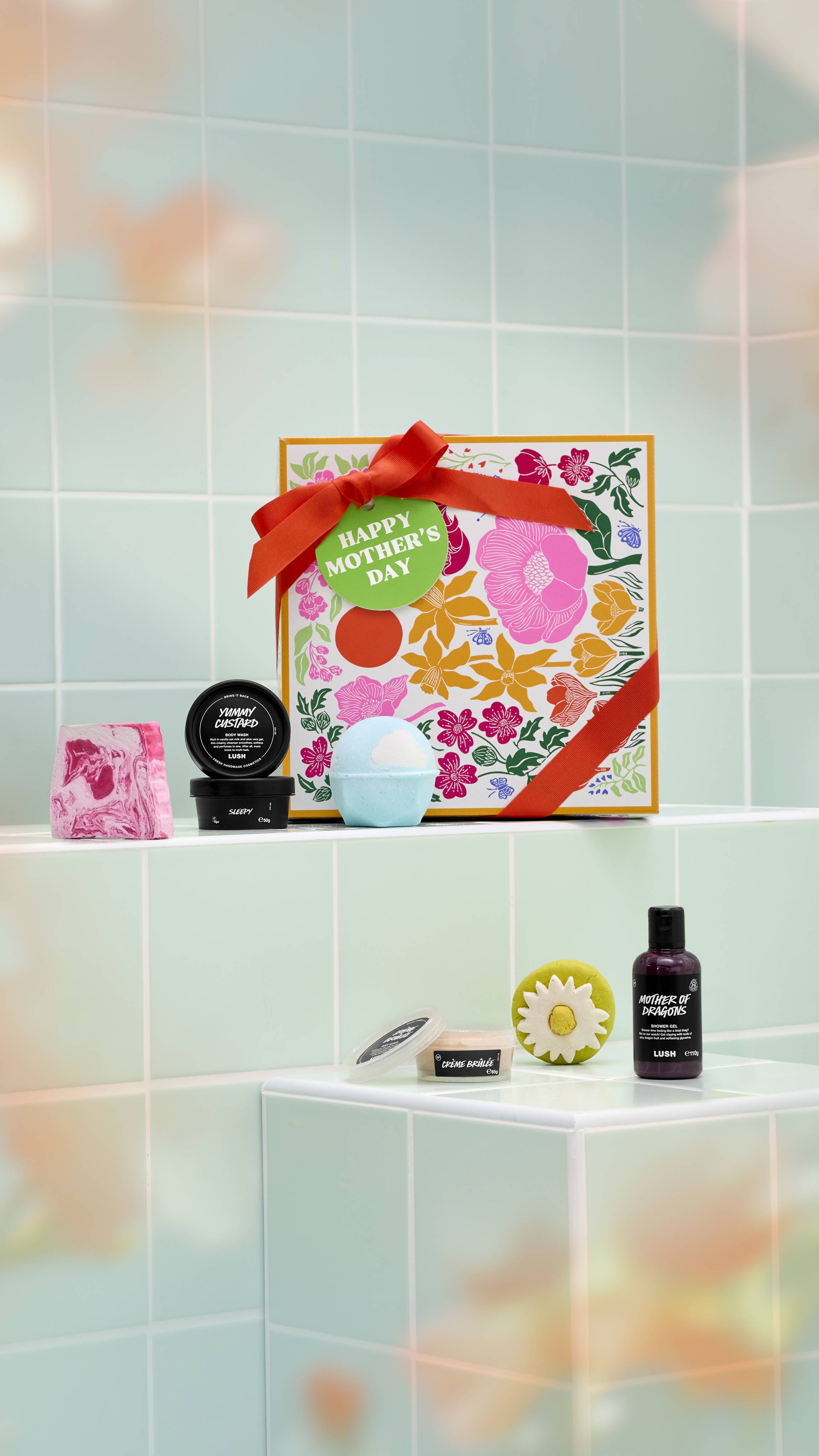 A square Lush "Happy Mother's Day" gift box sits on a pale green tiled ledge. Arranged around it are several bath and body products, including bath bombs, a soap, a bubble bar, and three potted products.