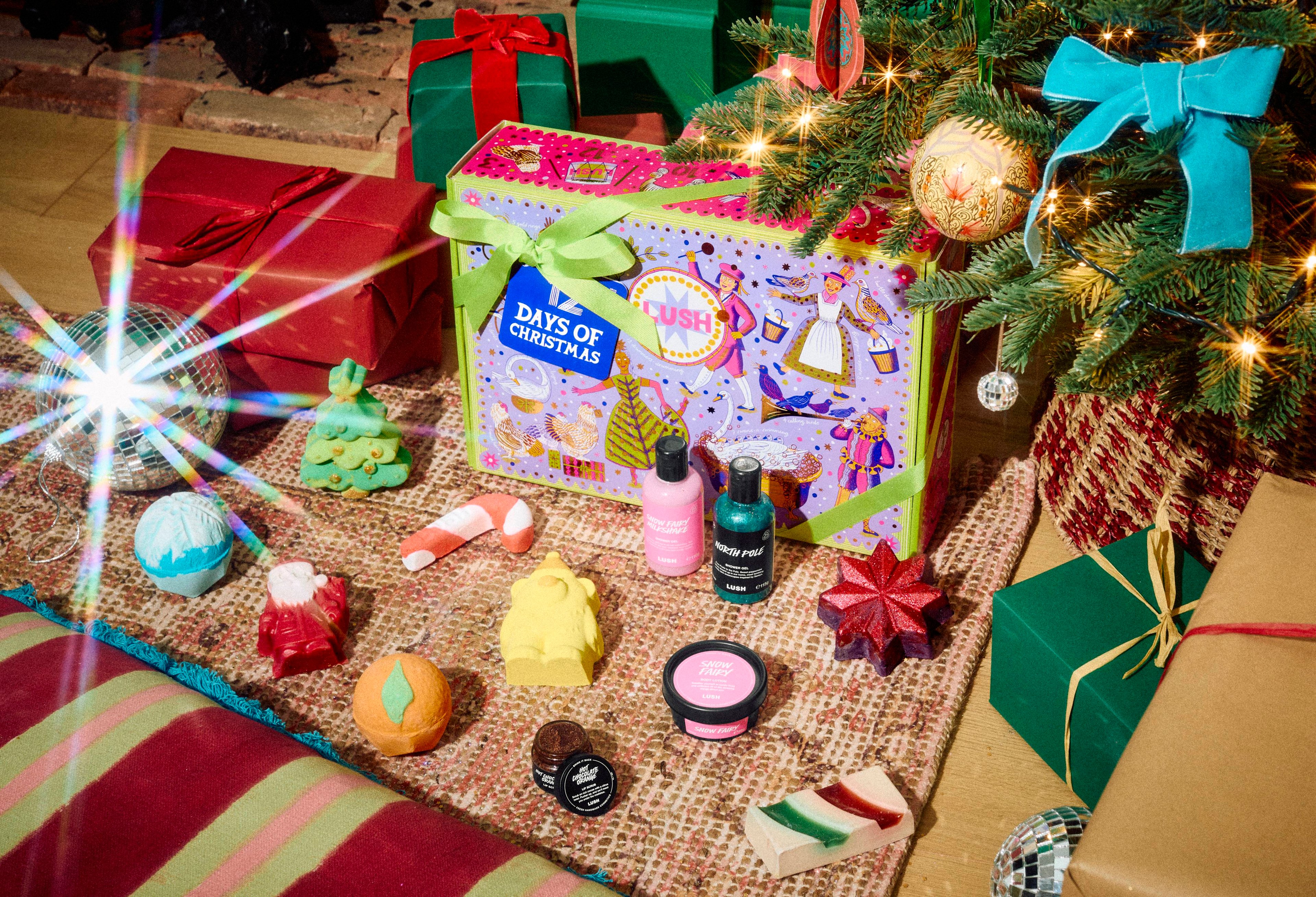 A colourful Lush '12 Days of Christmas' gift box under the tree, tied with a green ribbon and blue tag, surrounded by festive bath bombs, shower gels, soap, and lip scrub next to wrapped presents.