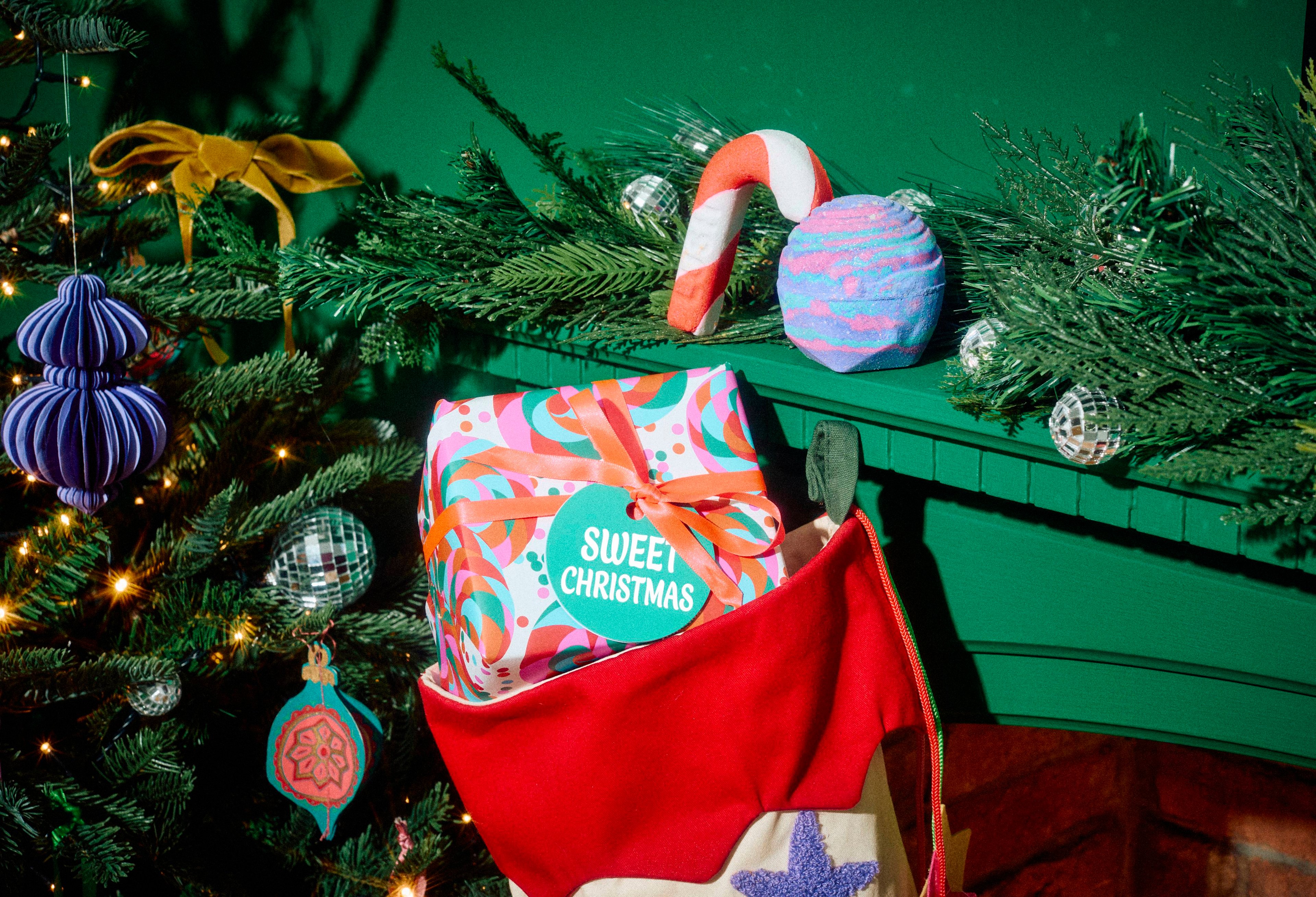 The Lush Sweet Christmas gift set tucked inside a red stocking, with a candy cane bubble bar and a purple swirl bath bomb displayed on a green mantelpiece surrounded by festive greenery.