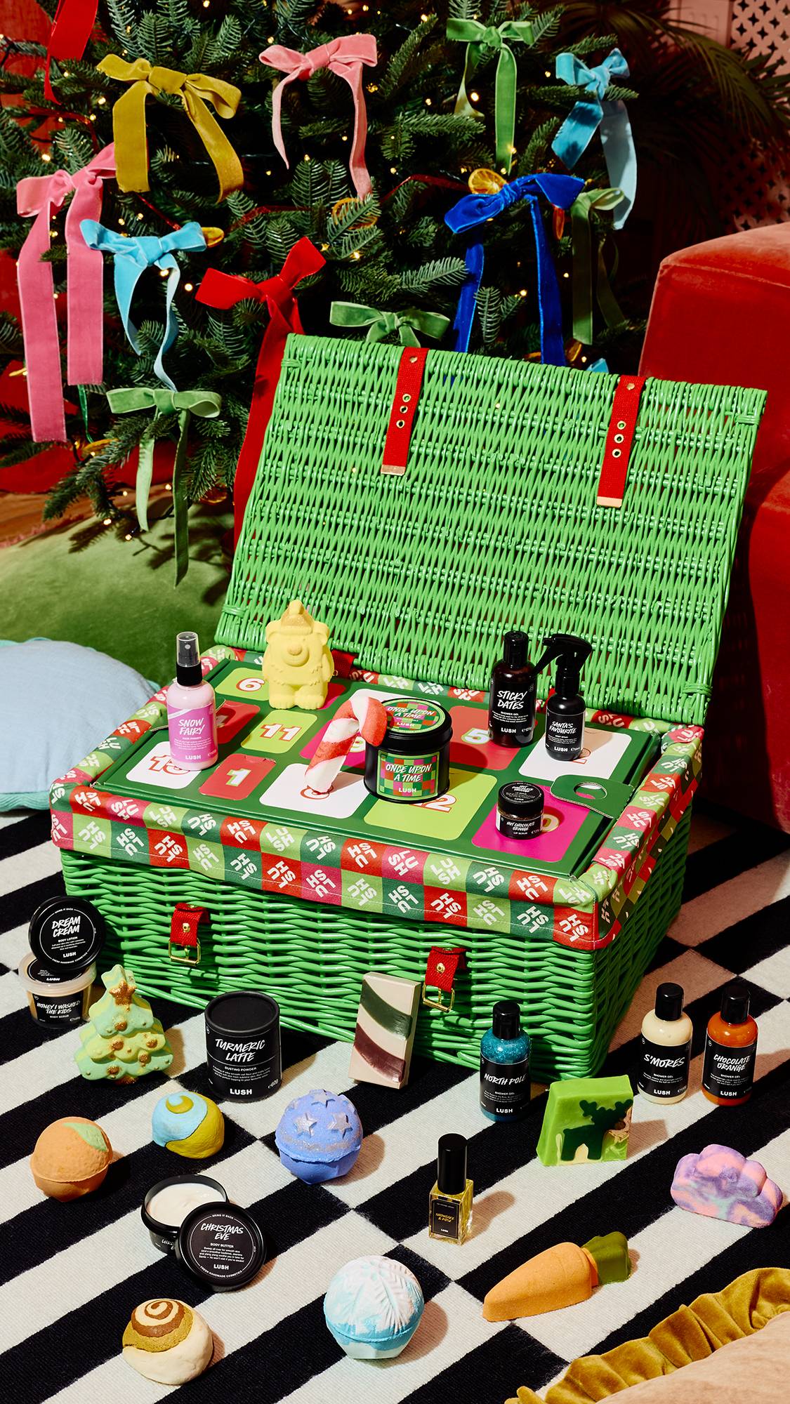 An open green hamper with red straps displays festive Lush treats, surrounded by Christmas bath bombs, soaps, and bottles scattered on a striped rug before a Christmas tree adorned with colourful bows.