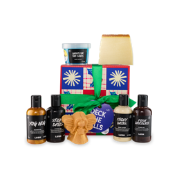 A colorful "Deck the Halls" gift box with a red and blue checkerboard pattern and snowflakes is tied with a green ribbon. Various Lush products are arranged around it, including shower gels, a body lotion, a body scrub, and two solid soaps.