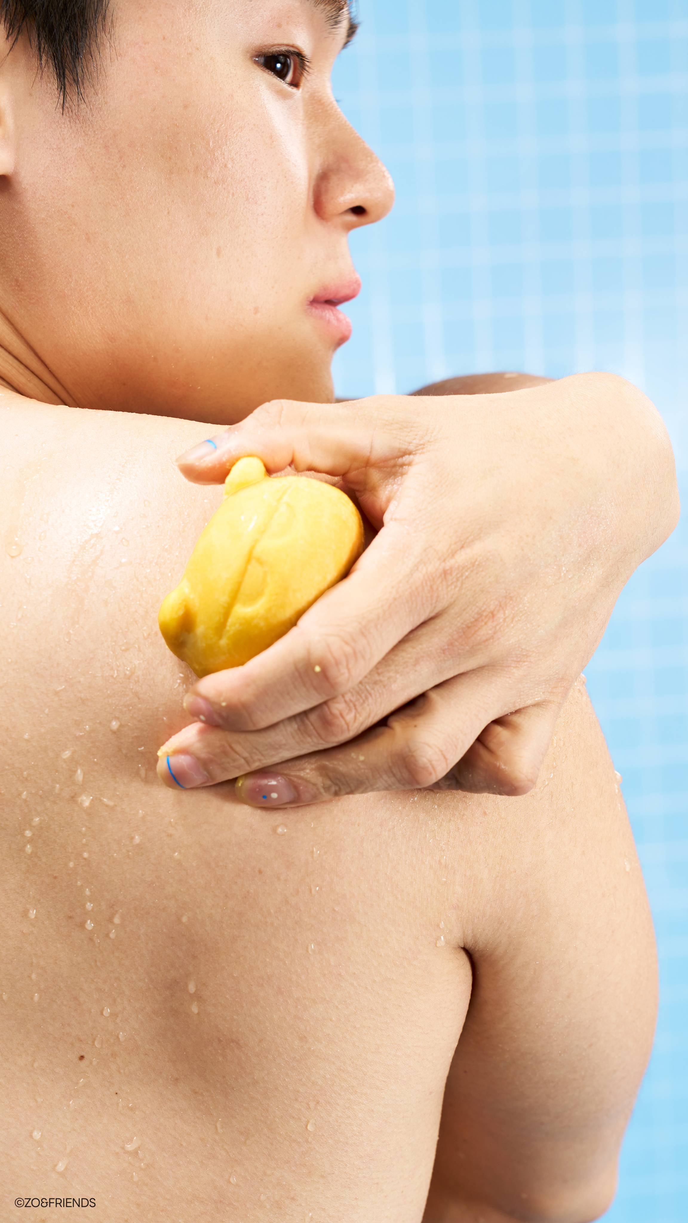 A close-up profile view of a person using the yellow Zaki character soap on their shoulder. Their wet skin is covered in tiny water droplets against a bright blue tiled background