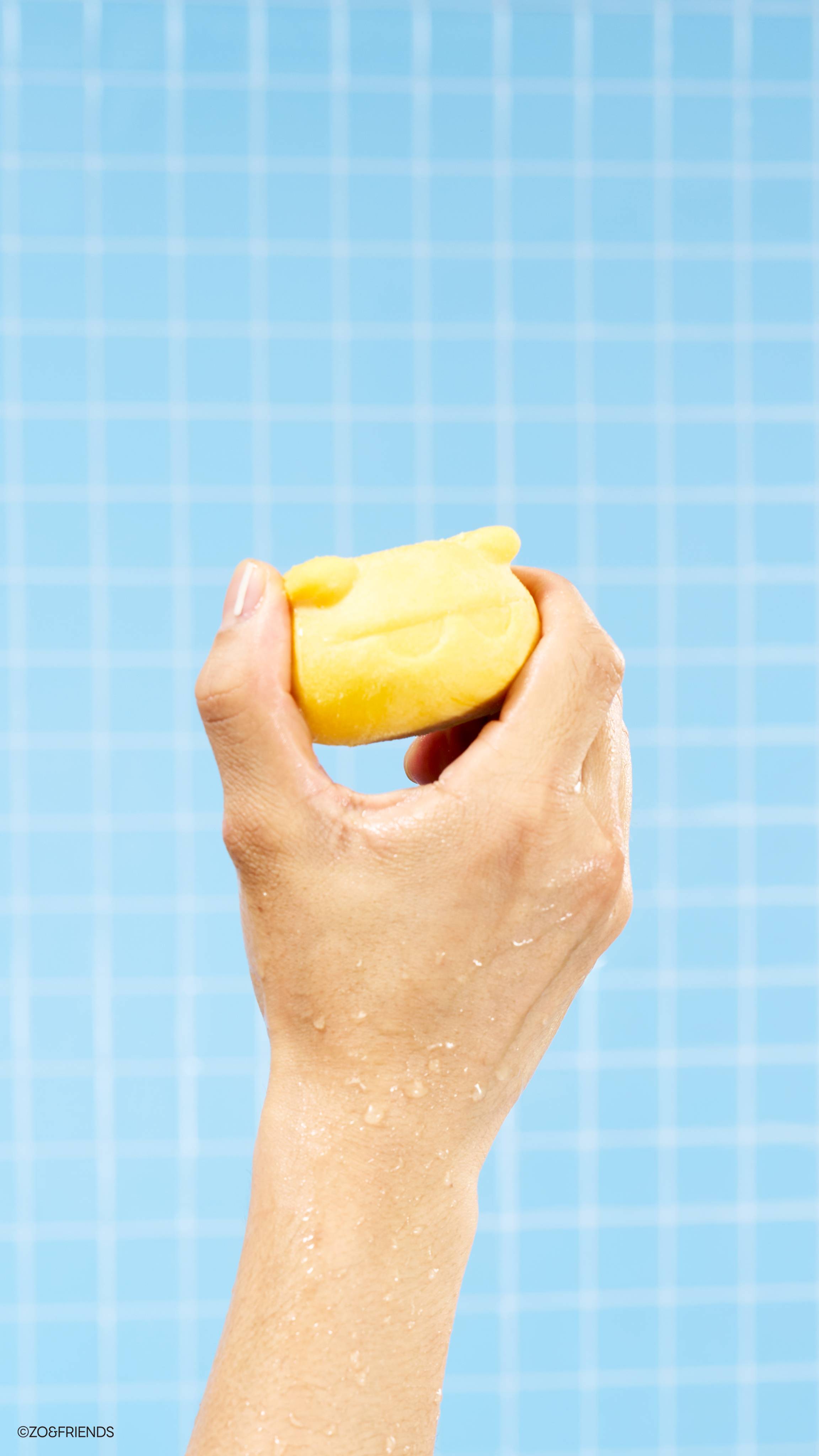 A wet hand covered in water droplets holds the yellow Zaki character soap up against a light blue tiled wall. The cat-shaped soap is held firmly between the thumb and fingers of an upright hand.