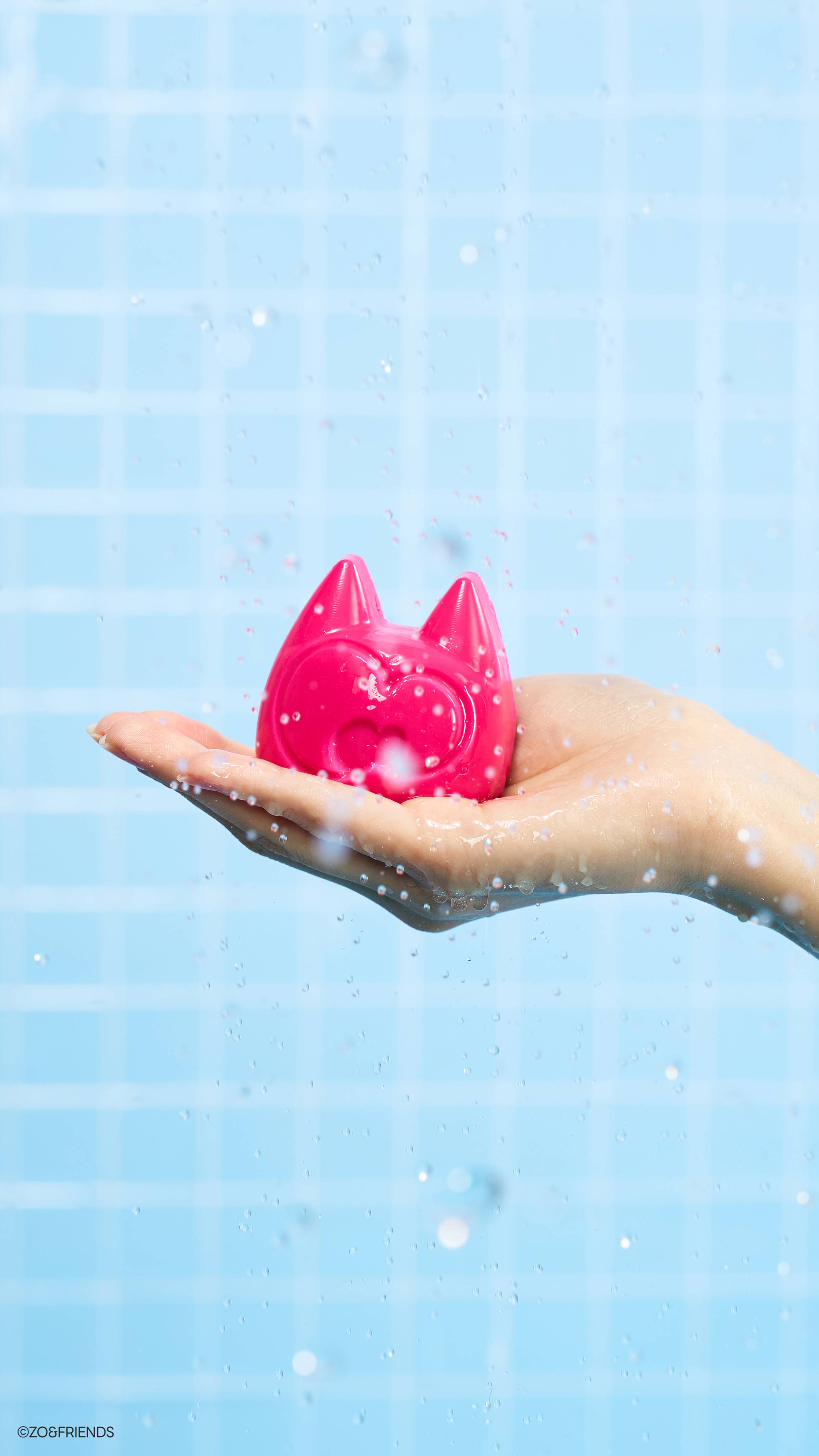 A hand holds the pink Aki character soap in an open palm under a spray of water droplets. The vibrant pink soap sits against a bright, light blue tiled background, captured mid-shower with splashing water frozen in the air
