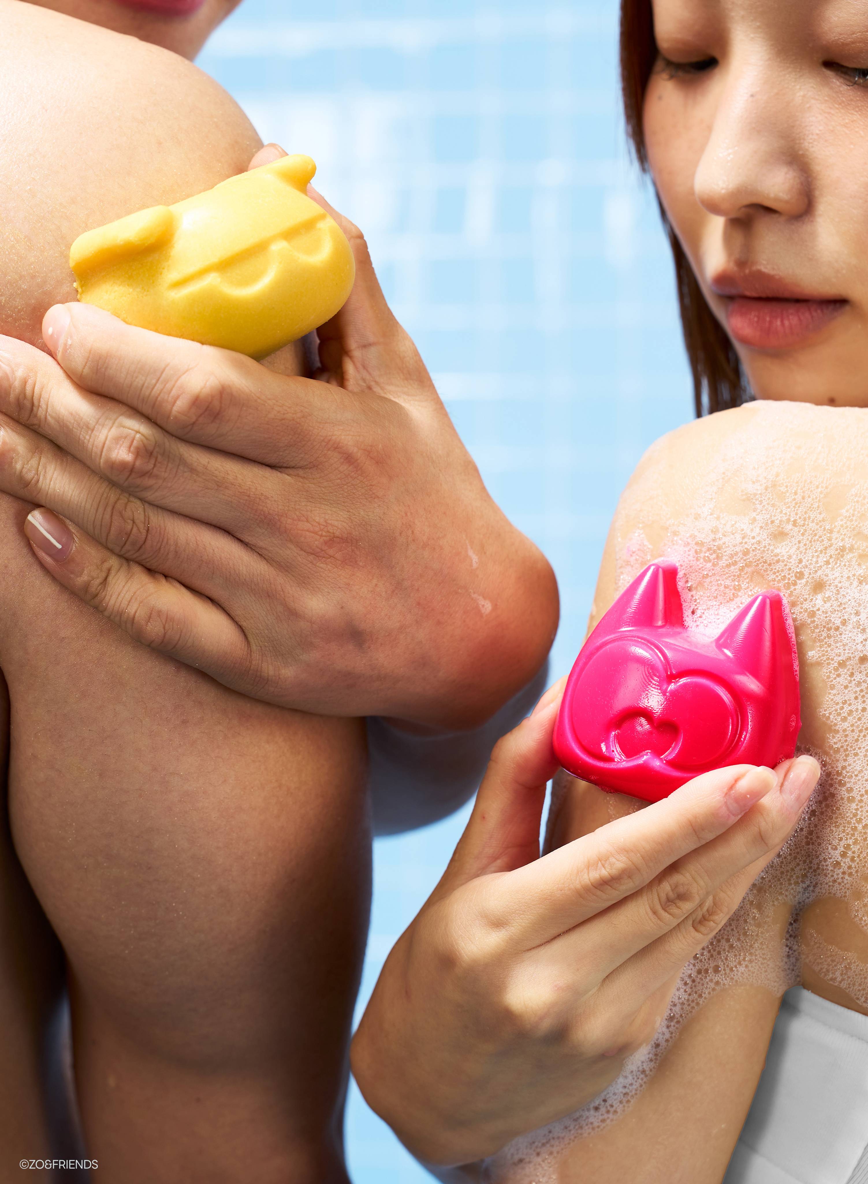Two people hold character-shaped soaps in a shower. One person holds the yellow Zaki soap against a bare shoulder. The other lathers the vibrant pink Aki soap onto soapy skin. They stand against a light blue tiled wall