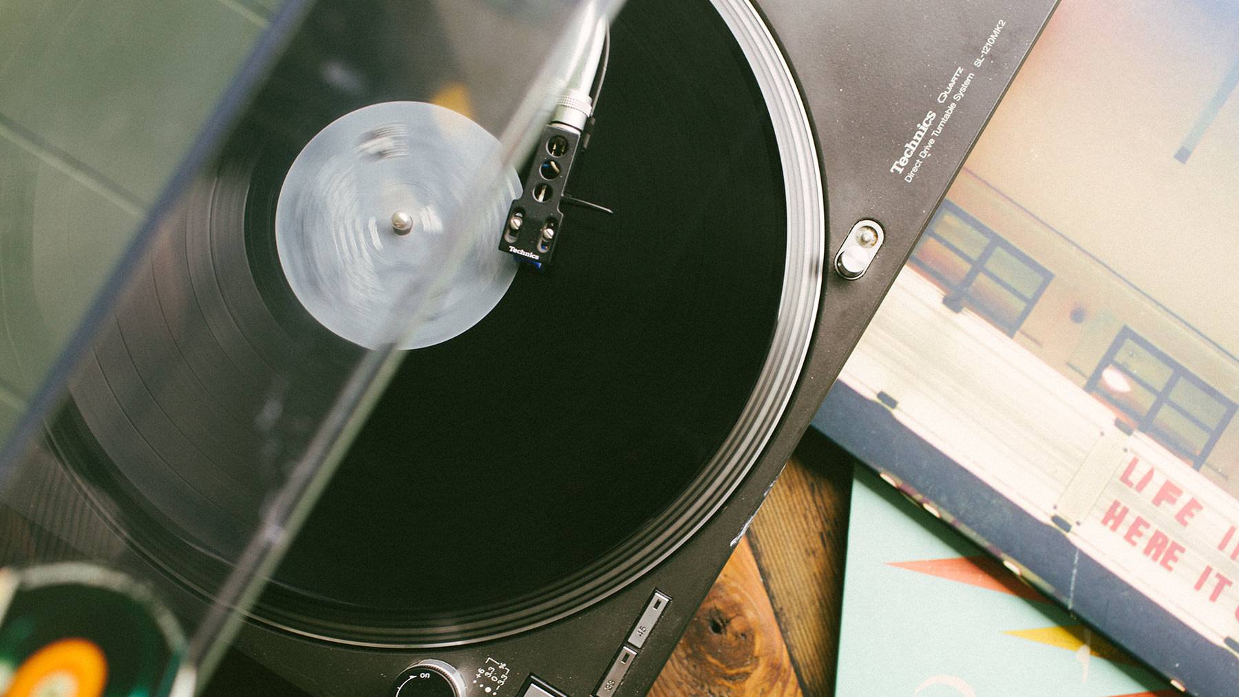 A high-angle, close-up shot of a Technics record player playing a black vinyl disc under a clear plastic lid, with vinyl record sleeves resting alongside on a wooden surface.