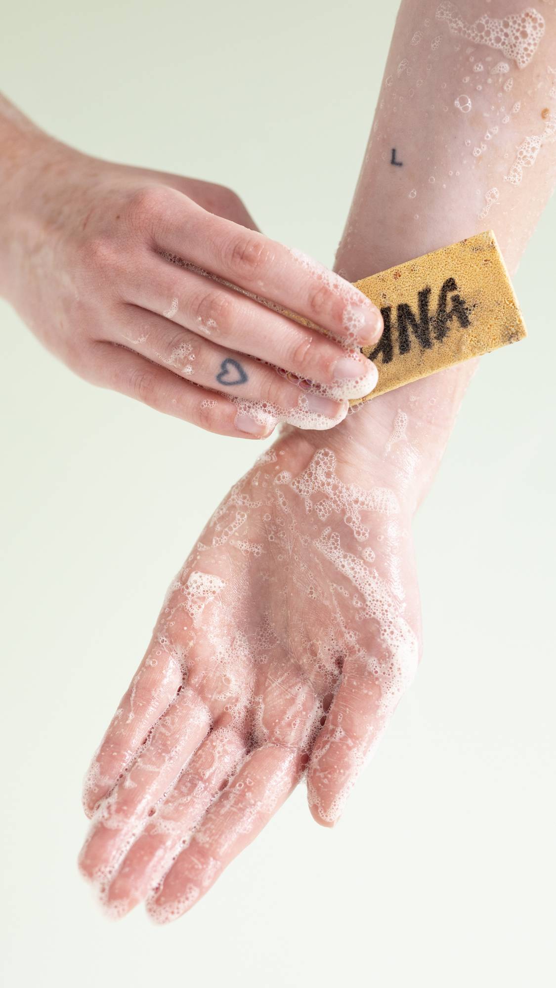 The image shows a close-up of the model's hands as the lather up the washcard under running water. 