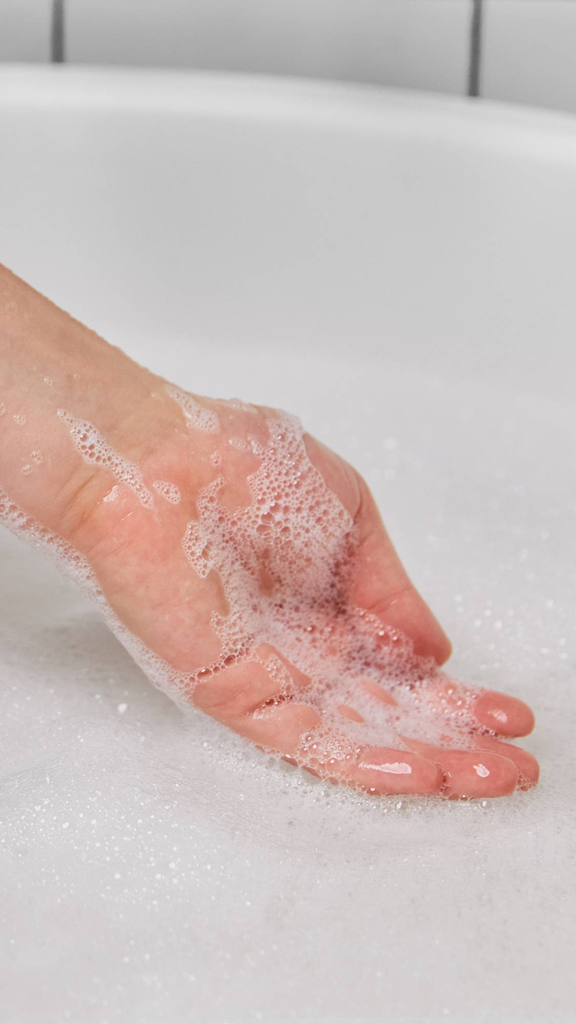 The image shows the model's hand after just having scooped up some thick, white bubbles from the bath water below. 