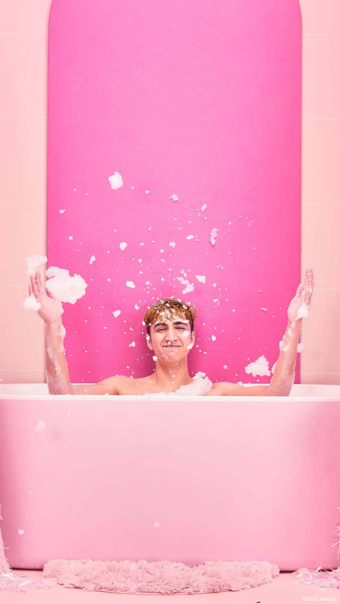 Image shows the model sat in the bathtub in front of a bright pink background after having just thrown bubbles in the air.