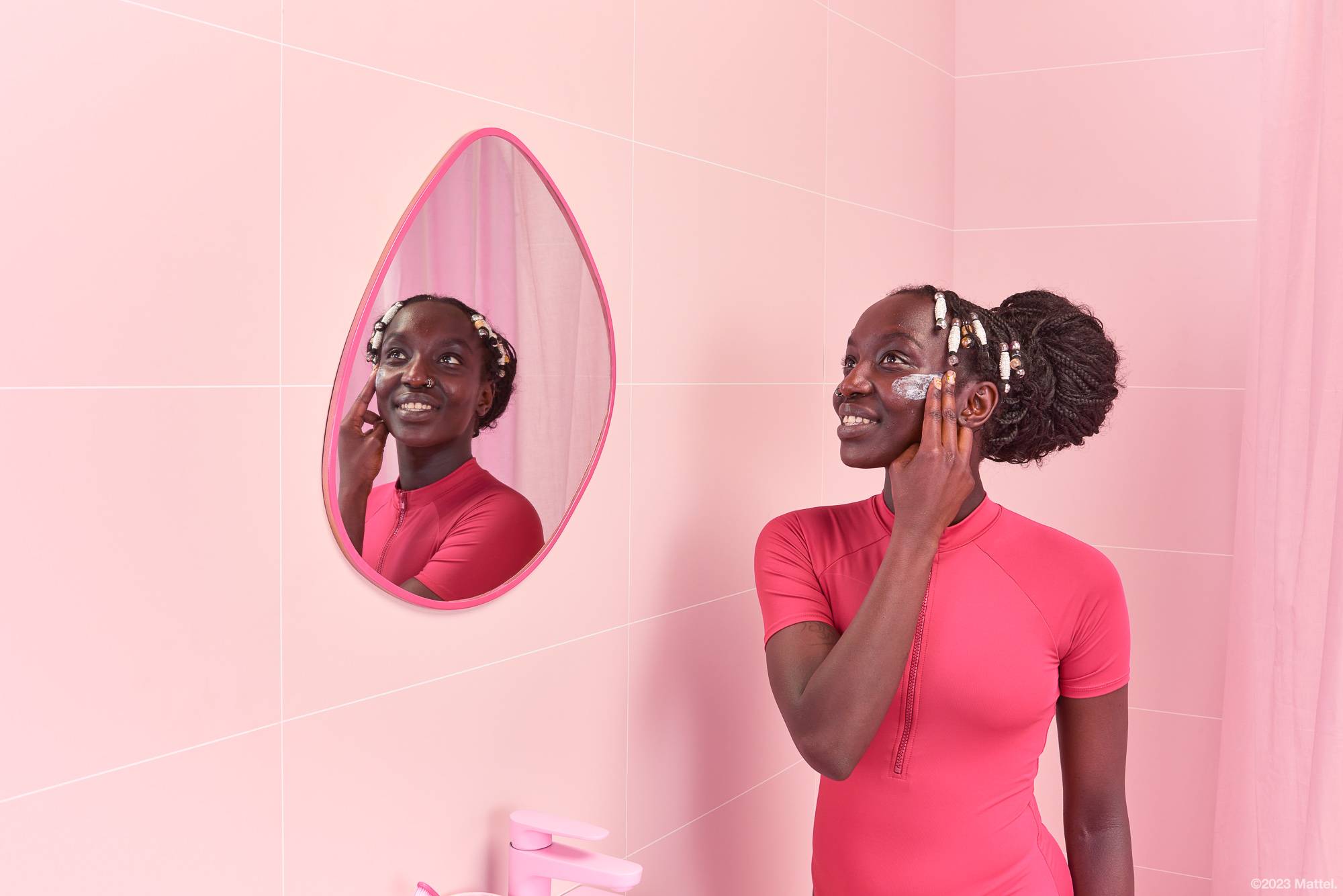 Image shows model in a pink bathroom with a mirror reflecting their face as they glaze their cheek with the product.