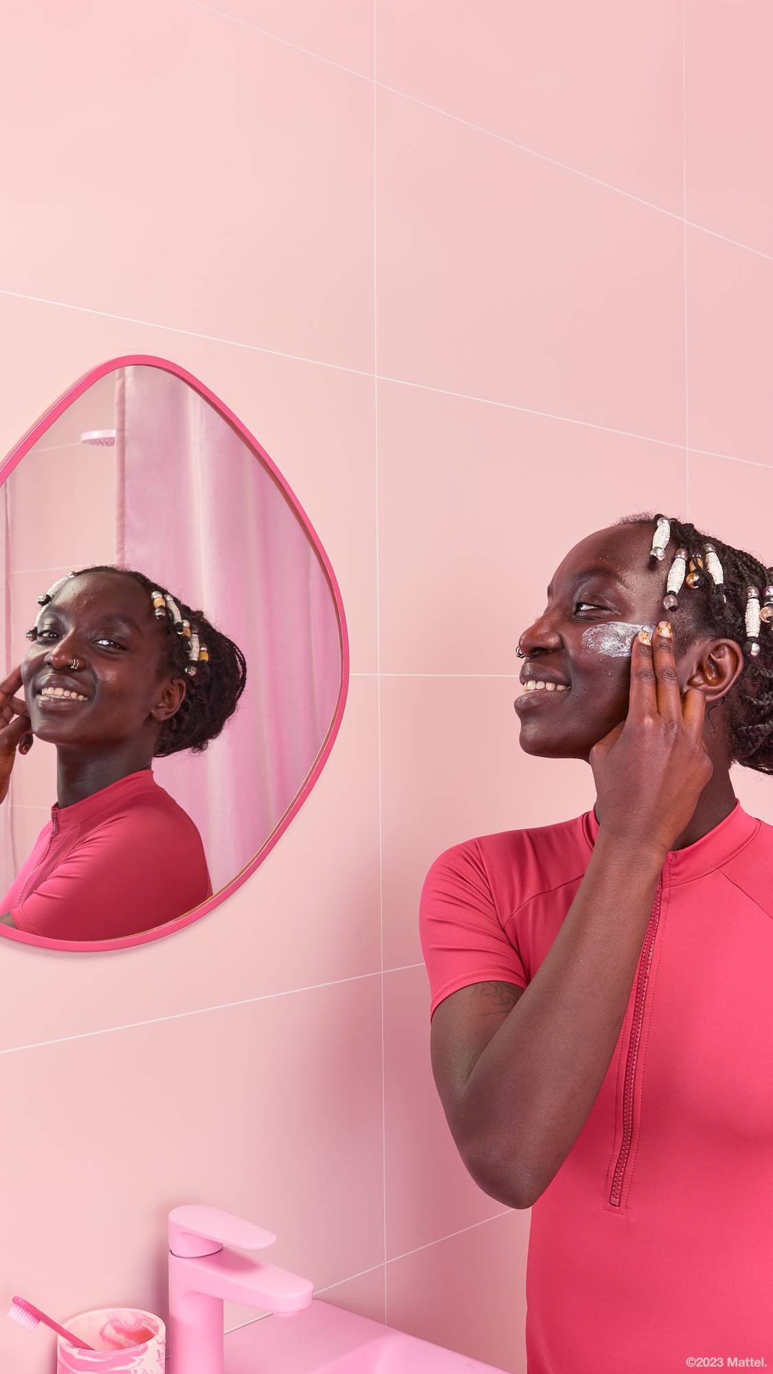 Image shows model in a pink bathroom with a mirror reflecting their face as they glaze their cheek with the product.