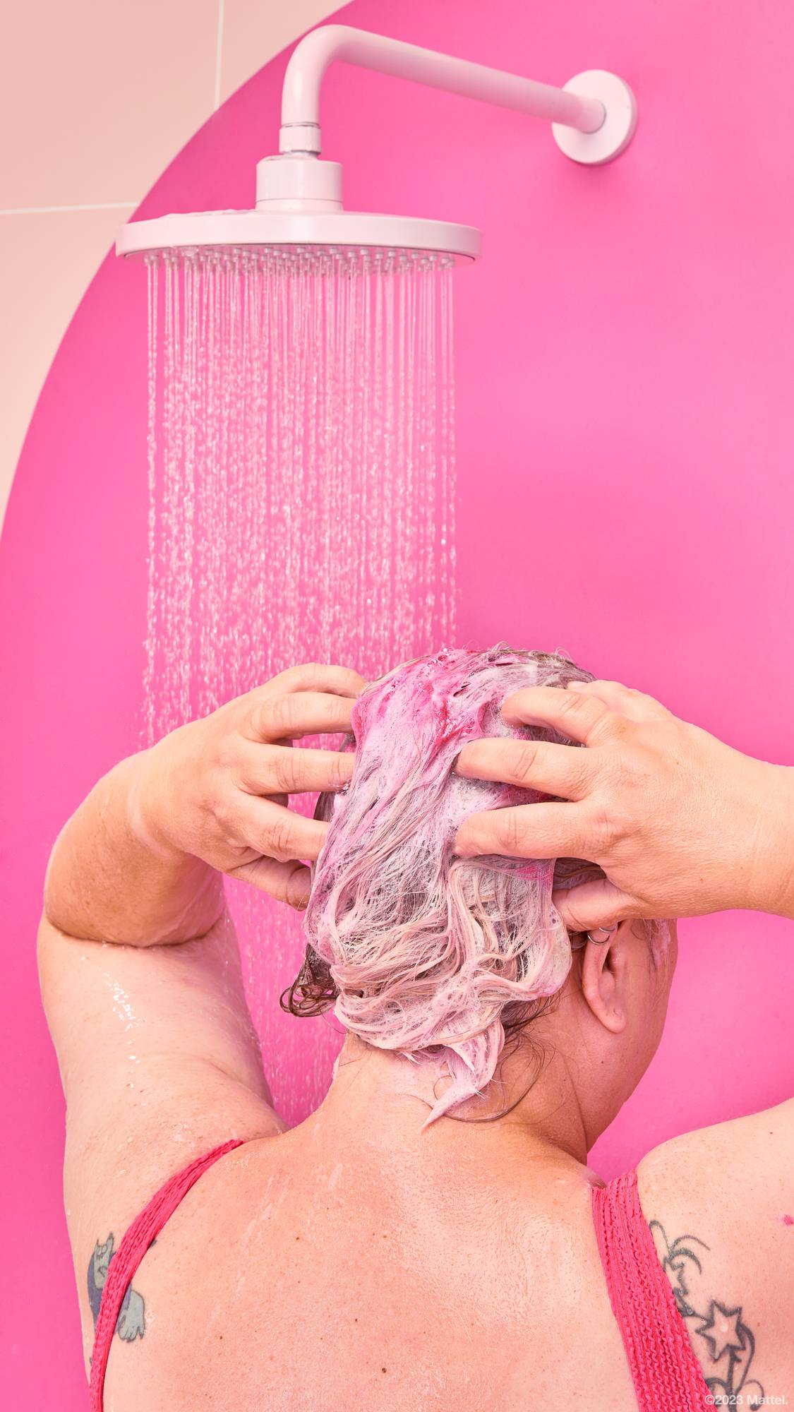 Image shows the model as they stand under the shower water. They are lathering up their hair which has a subtle pink tint. 