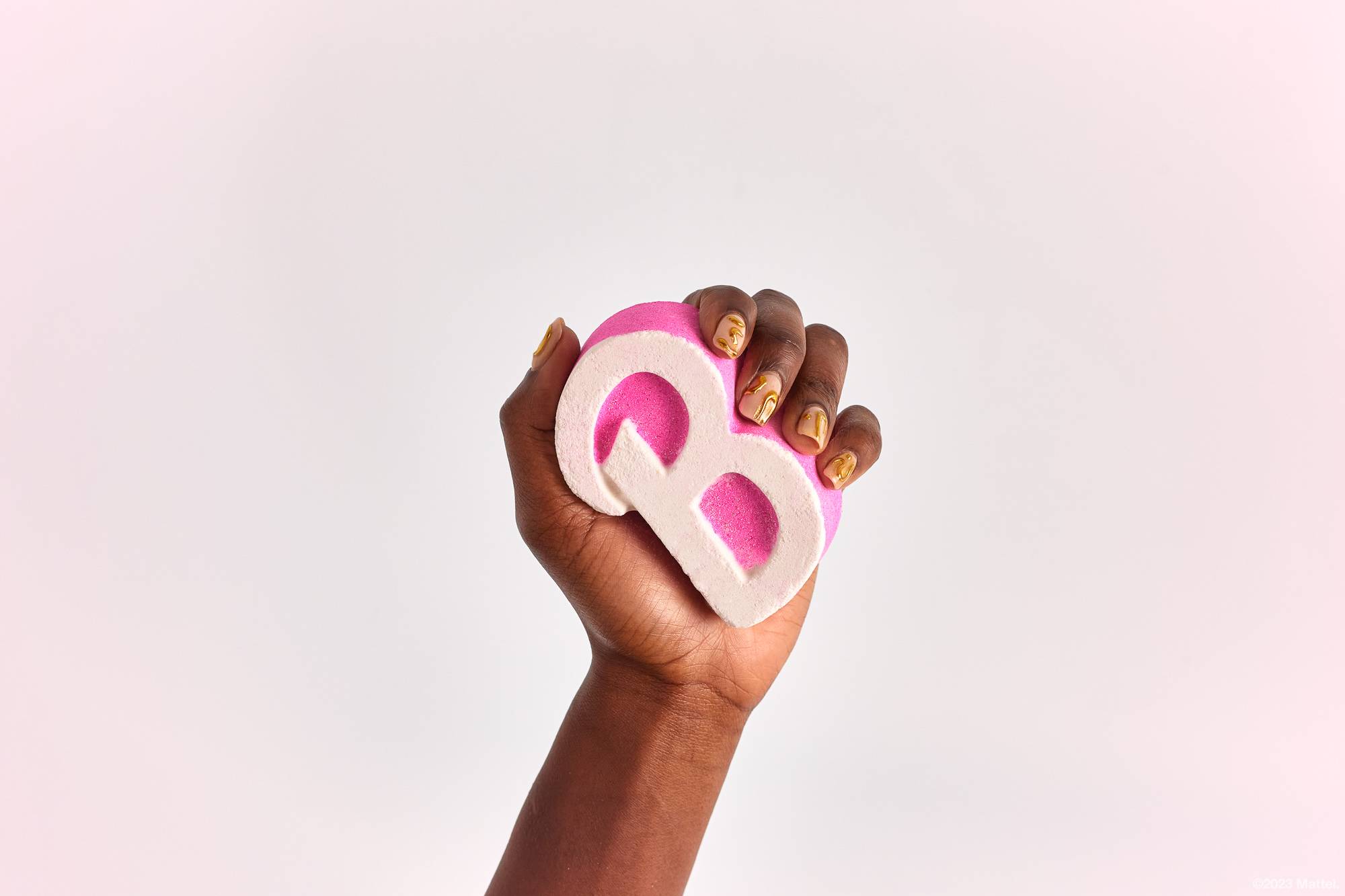 Image shows a close-up of the model's hand holding the Barbie bath bomb in the air in front of a solid white background. 