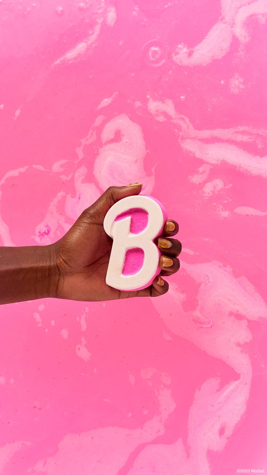 Image shows close-up of model's hand holding the Barbie bath bomb above a background of swirling pink bath water. 