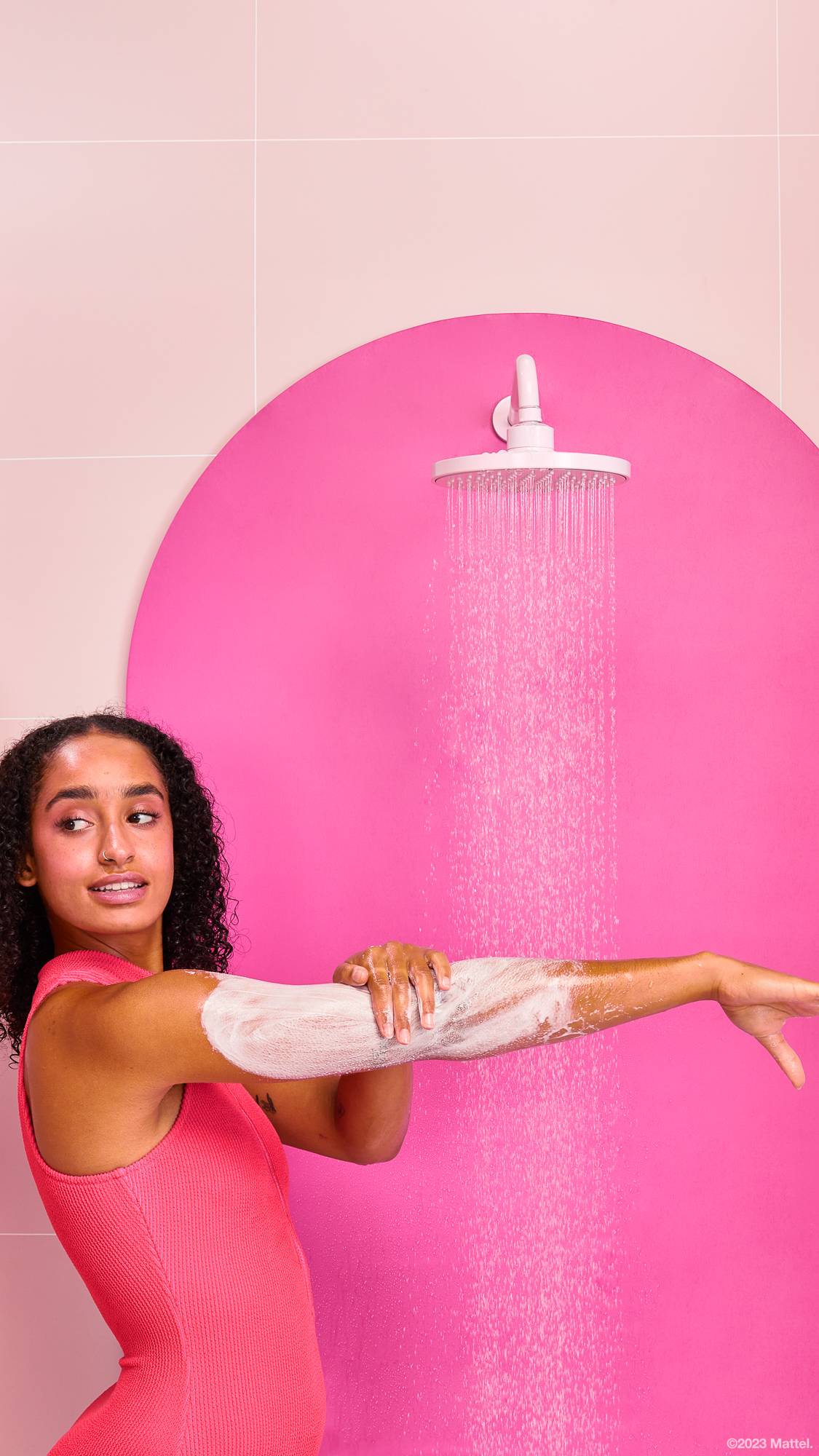 Model is stood under a running shower in a pink swimming costume. They are exfoliating the body scrub over their arm.