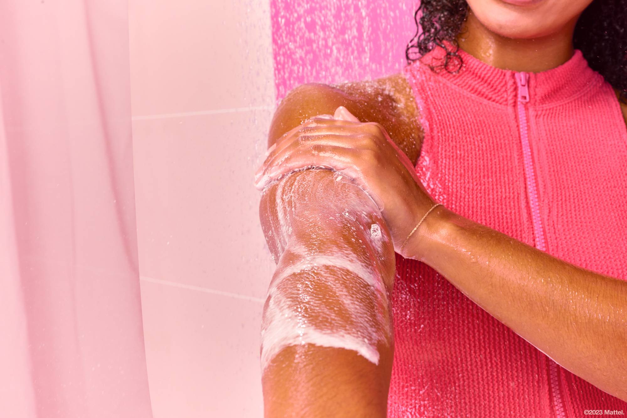 Close-up of model's arm under running shower water as the model exfoliates the Barbie Dream body scrub over their skin.