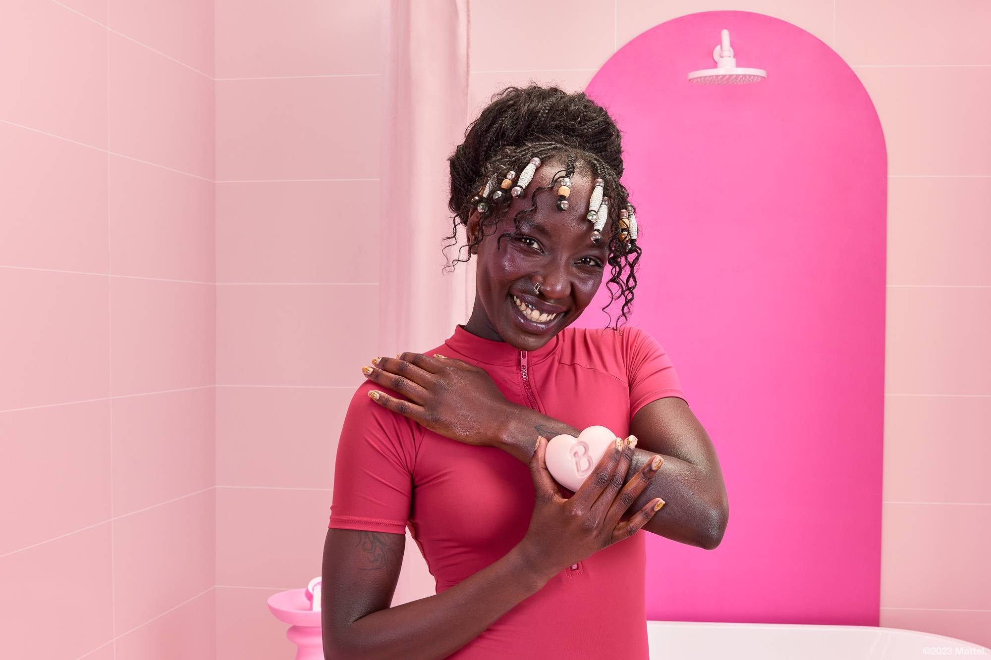 Model is in a pink bathroom. They are wearing a pink swimming costume and smiling as they soothe the body balm over their arm.