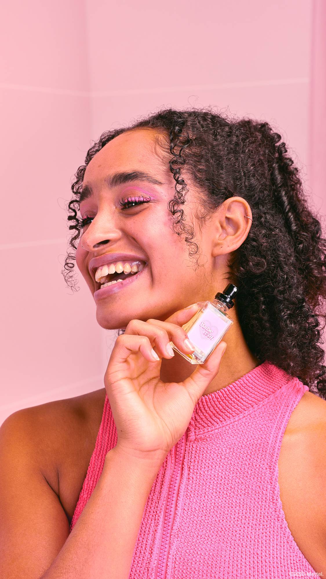 Model is holding the perfume bottle up by their face. They are smiling, wearing pink make up and clothing on a pink background.