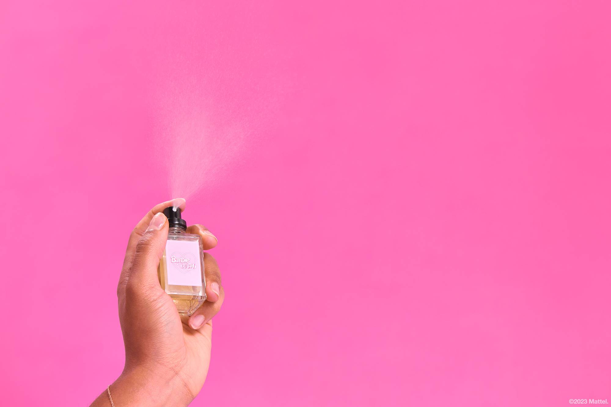 An outstretched arm is holding the perfume bottle and gently spritzing in front of a bright pink background.