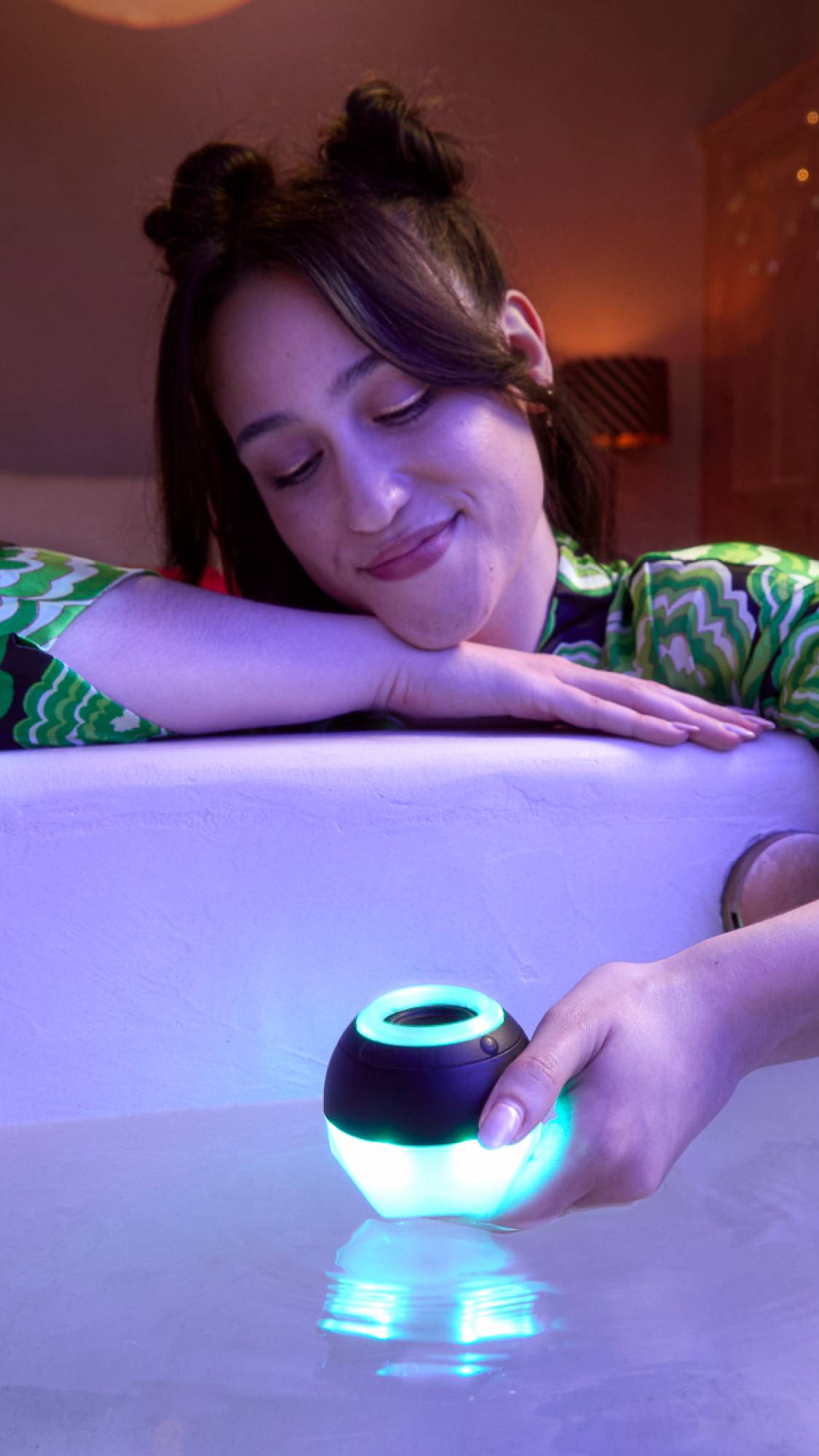 The model is sat over the bath with one hand lying on the side and the other holding the Bath Bot above the water. The Bath Bot is illuminated blue on the bottom half and around a small ring at the top. 