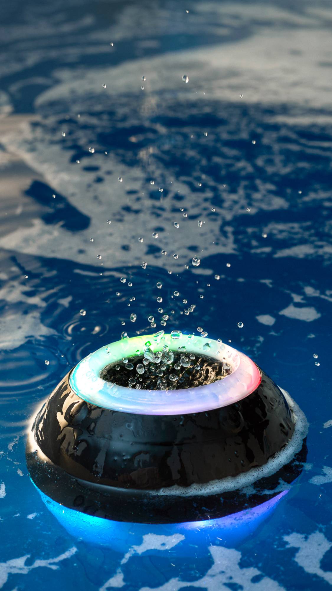 The black Bath Bot is lying in clear, deep blue waters. The upper ring light is illuminated rainbow as a puddle of water splashes from the speaker vibrations on top.