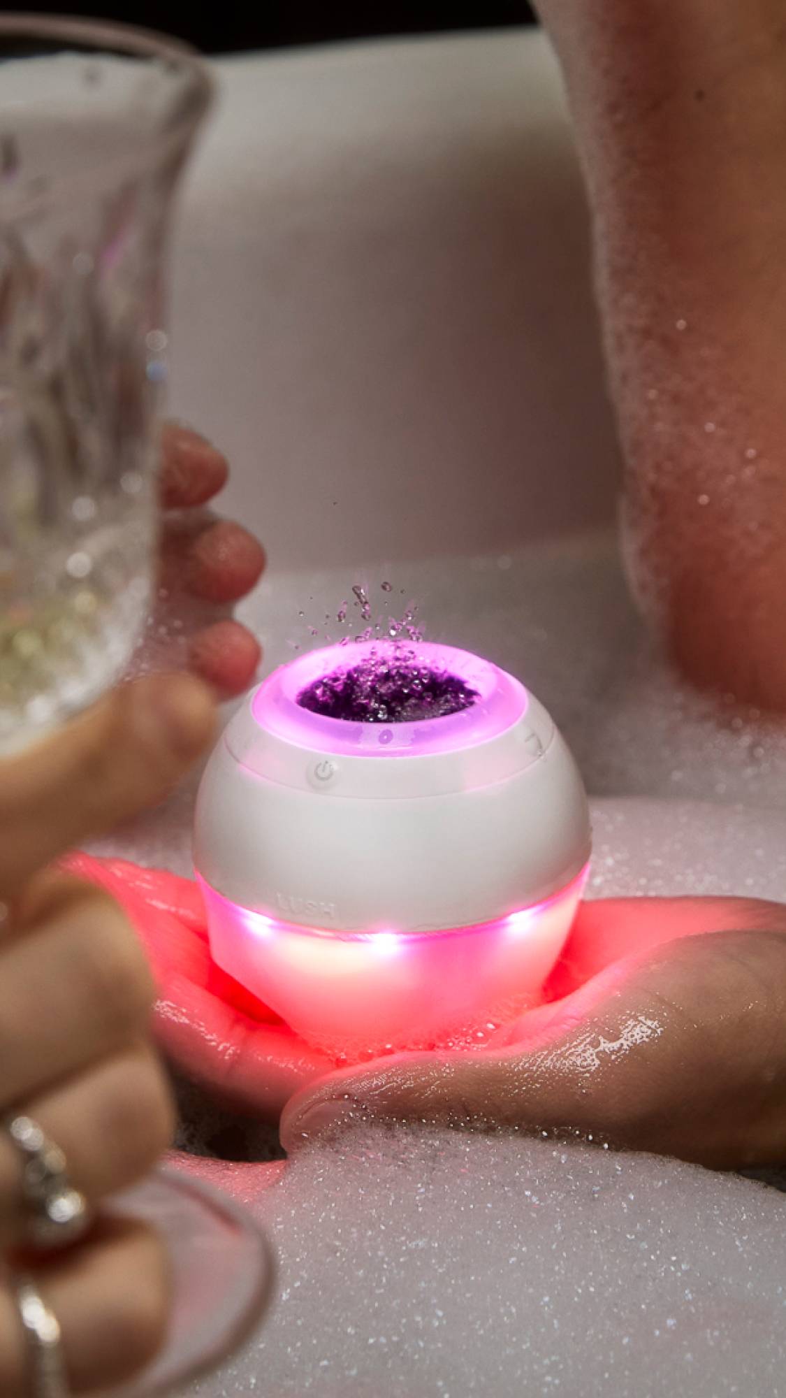 The model holds the Bath Bot in their hand above a very bubbly bath. The lower half of the Bath Bot is glowing a deep red-pink colour. The upper ring is glowing pink as droplets splash from the speaker. 