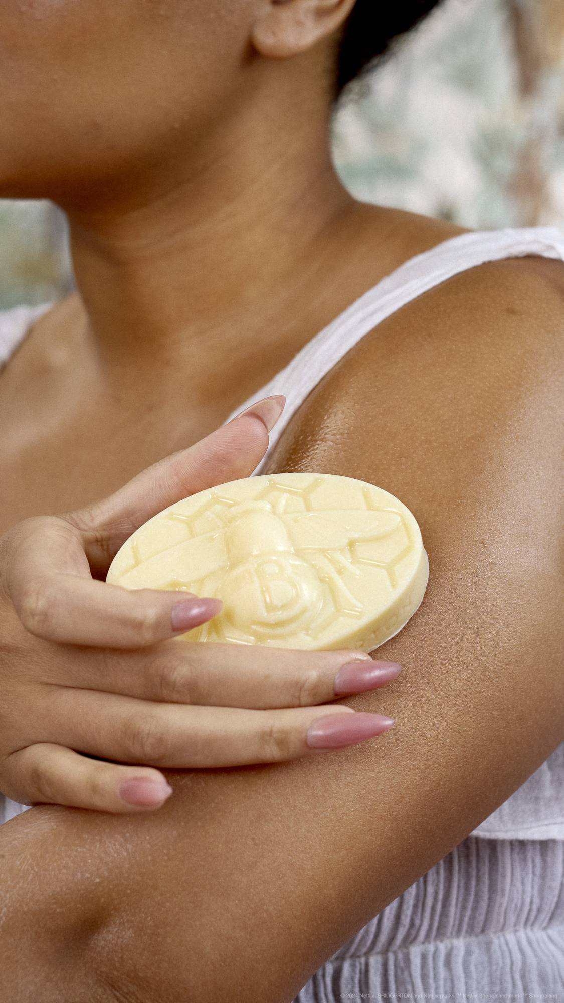 A close-up of the model's upper arm focuses on the Bee solid body balm as they apply it over their shoulder.